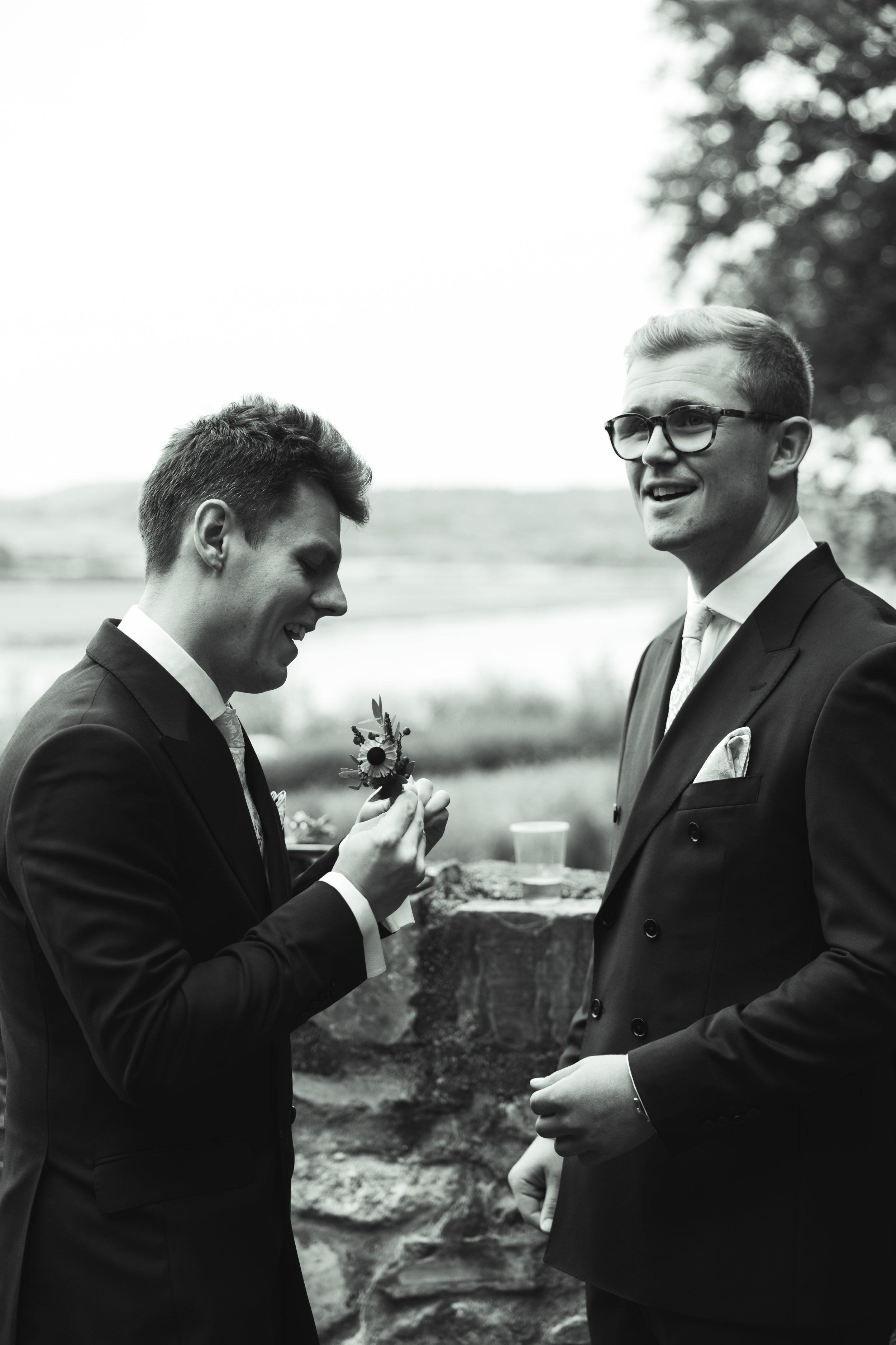 Black and white photo of two men in suits outdoors, one holding a small flower, smiling, with a blurred natural background.