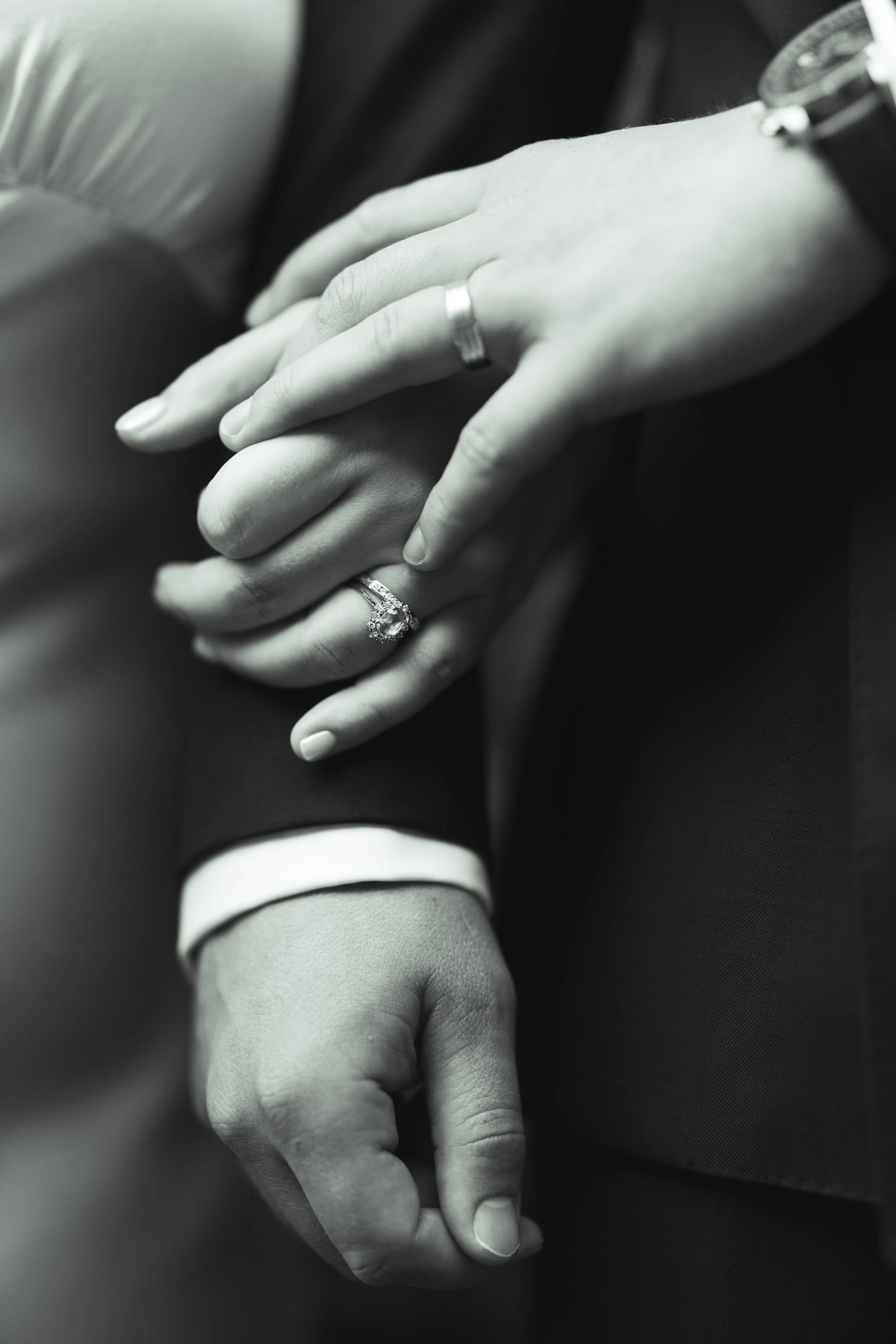 Close-up of two hands holding each other, one with a wedding ring and engagement ring, the other with a wedding band, in black and white.