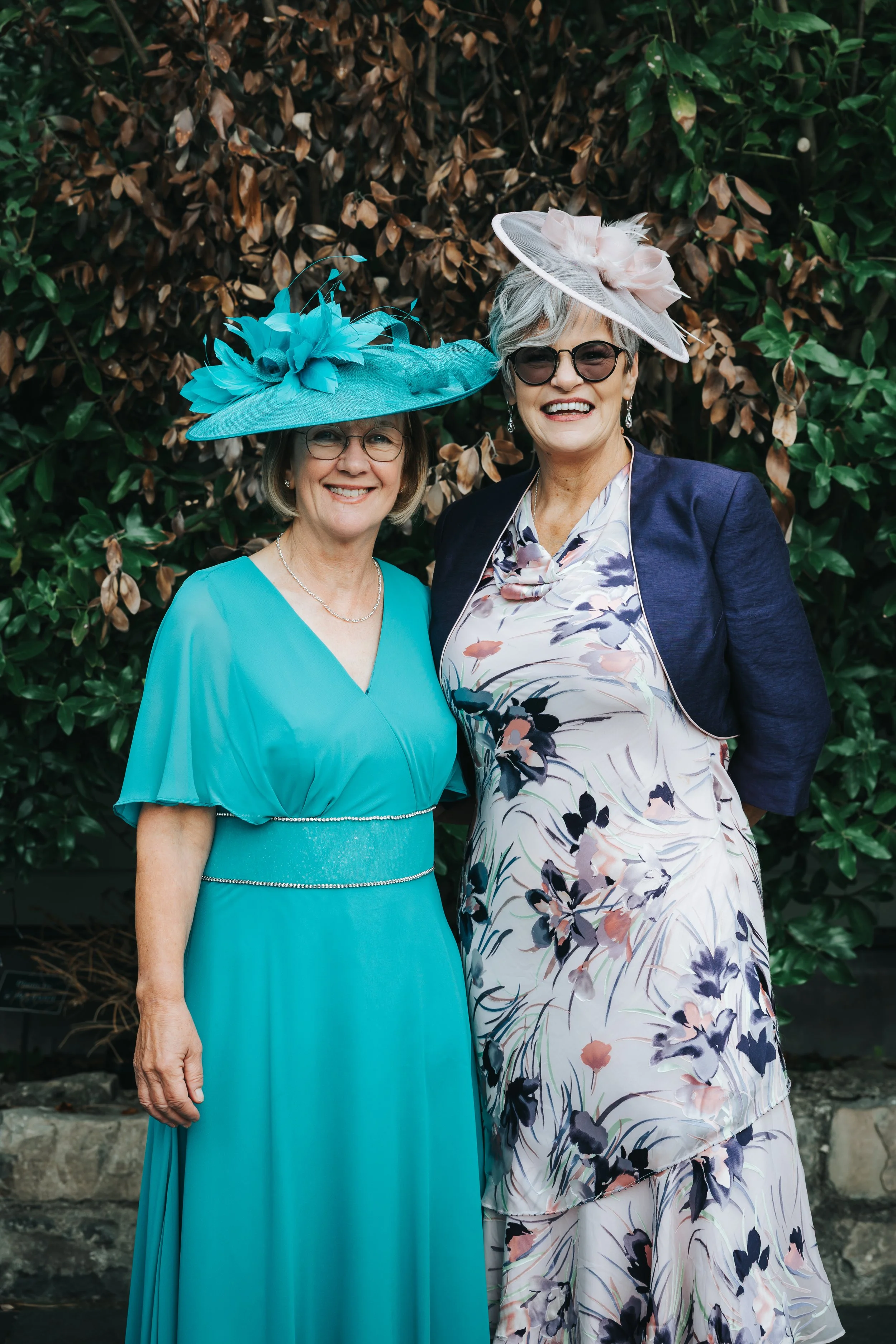 Two women standing outdoors, dressed in elegant dresses and large feathered hats, smiling at the camera.