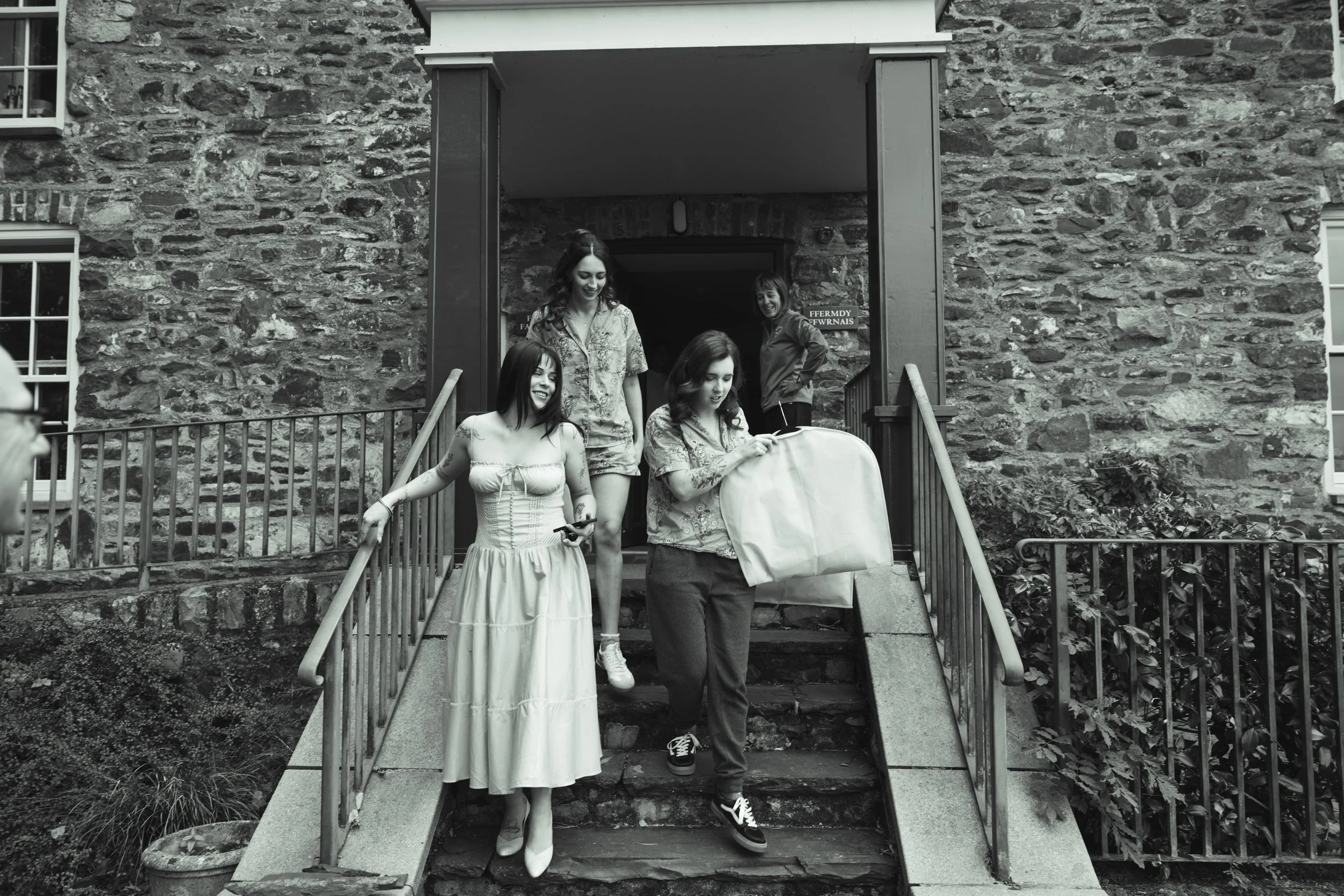 Four women descending stone steps outside a brick building, with a woman in the background, one holding a large bag.