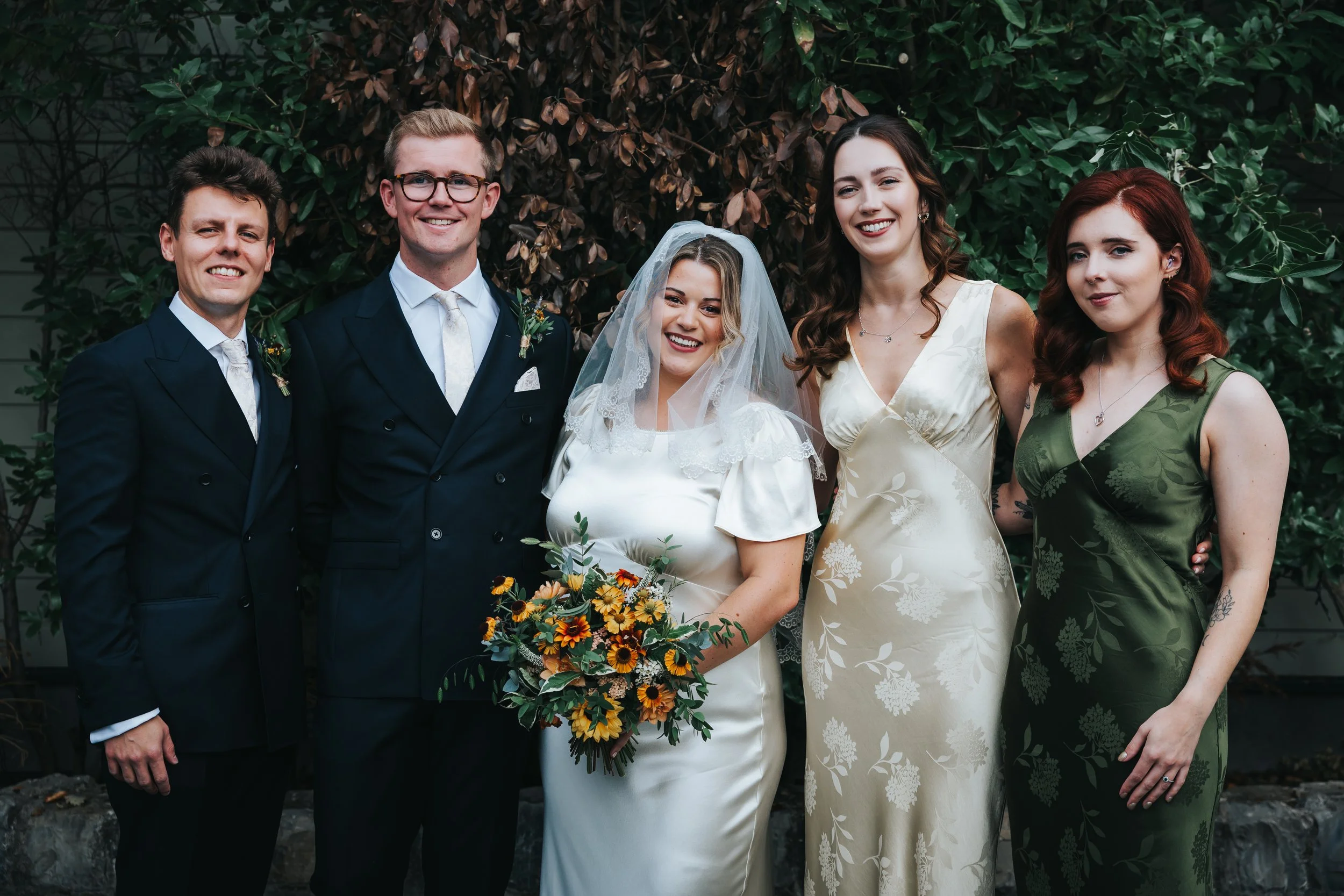 Group of six people at a wedding, including the bride in a white dress with a floral bouquet, standing alongside five friends or family members dressed formally, outdoors with leafy background.