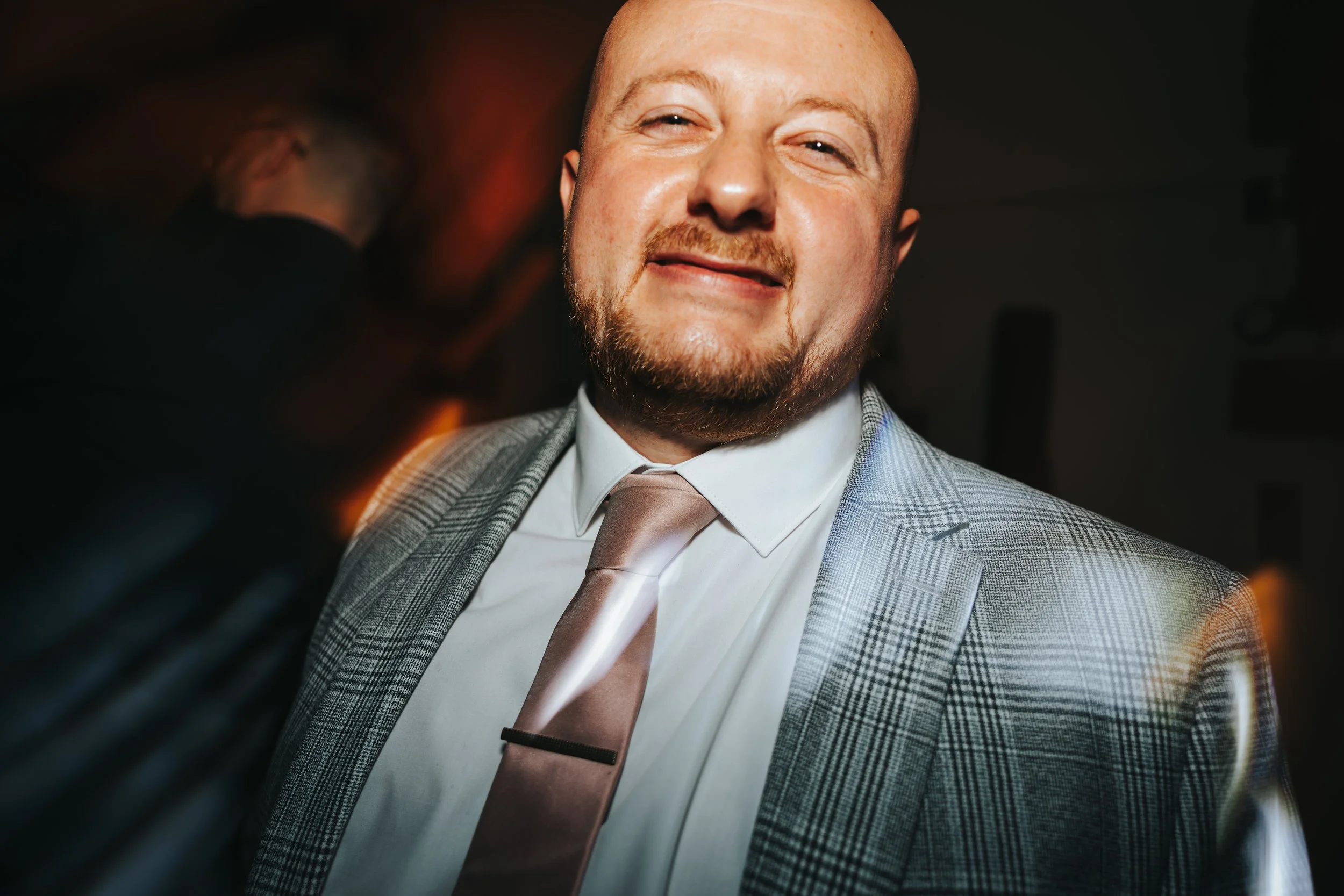 A man with a beard smiling and wearing a gray plaid suit, white shirt, and tan tie, standing in a dimly lit room.