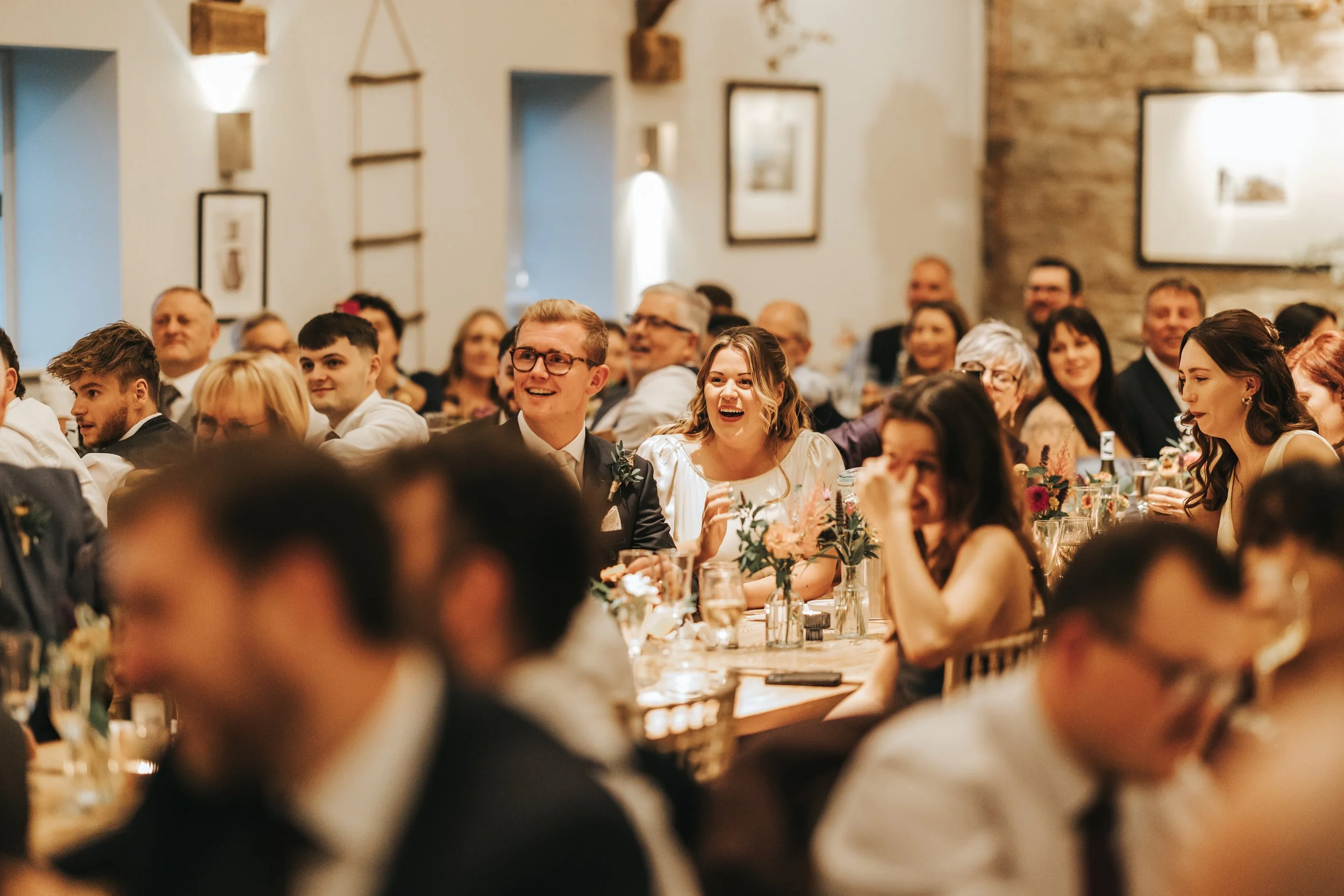 A group of people at a wedding reception, sitting at tables with floral centerpieces, laughing and smiling.