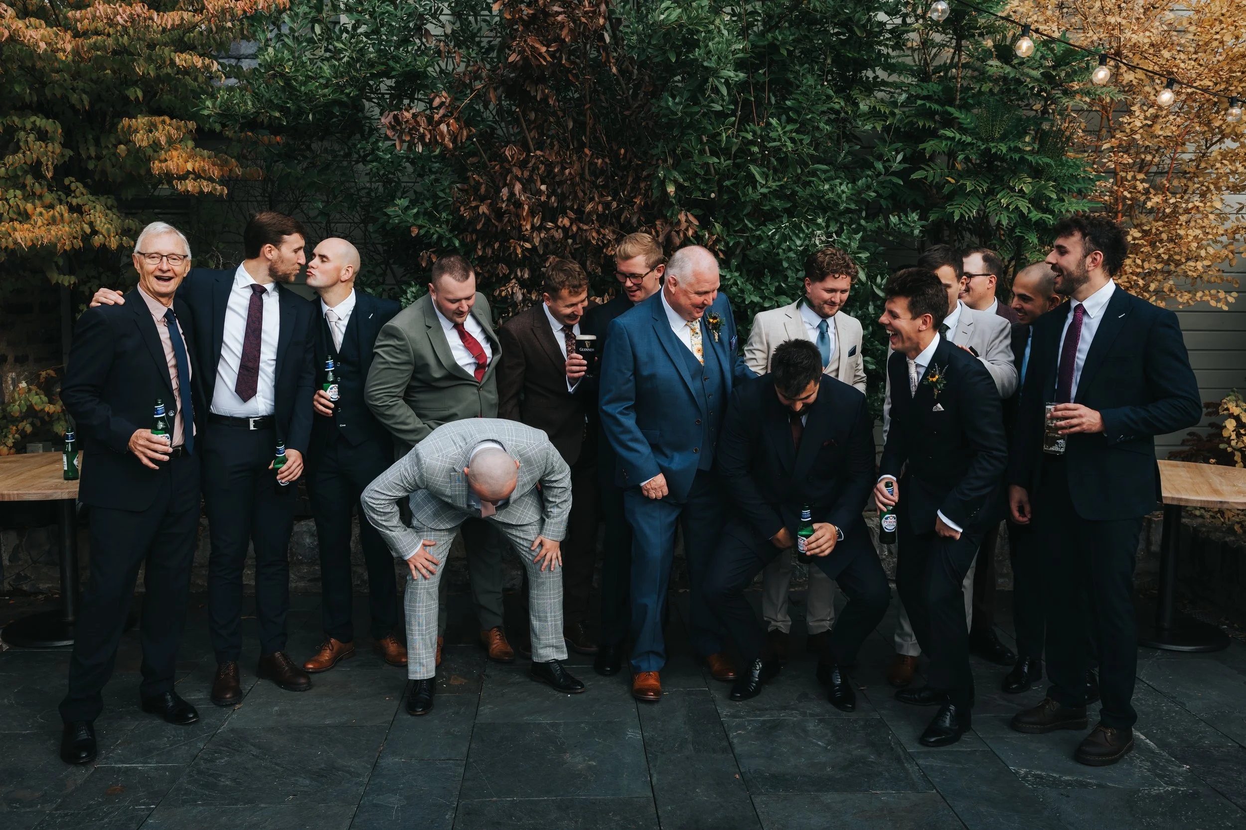 Group of men in suits at a wedding or formal event, standing outdoors against a backdrop of trees and string lights, some holding drinks, with one man bowing or bending down.