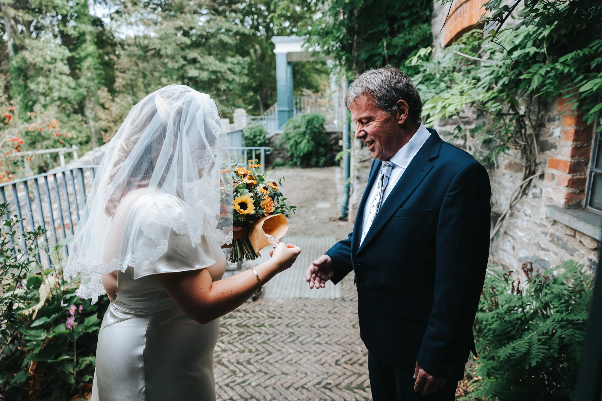 A bride in a white wedding dress and veil giving a flower to a smiling man in a suit outdoors, with greenery and a stone wall in the background.