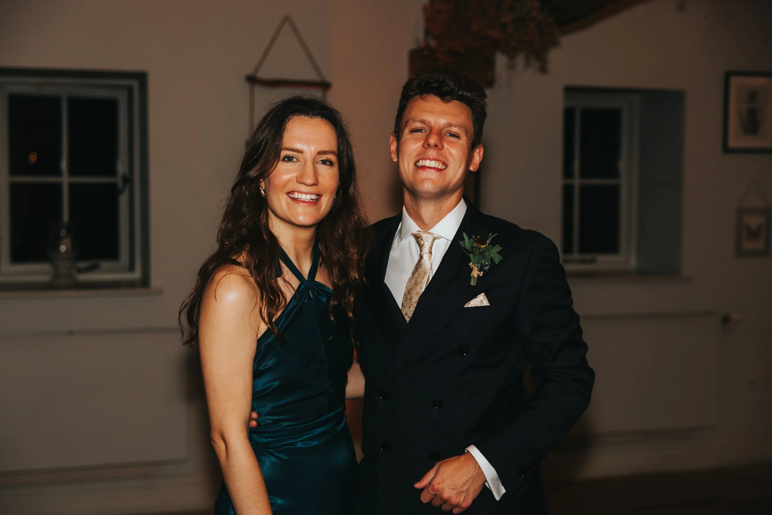 A smiling couple dressed in formal attire at a social event or celebration, standing indoors in front of a neutral-colored wall with windows.