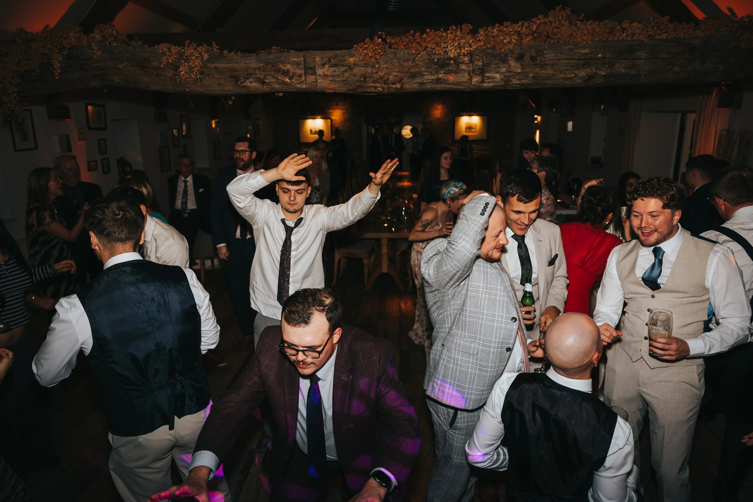 People dancing and socializing at a lively indoor event or party, with a wooden ceiling and framed pictures on the walls.