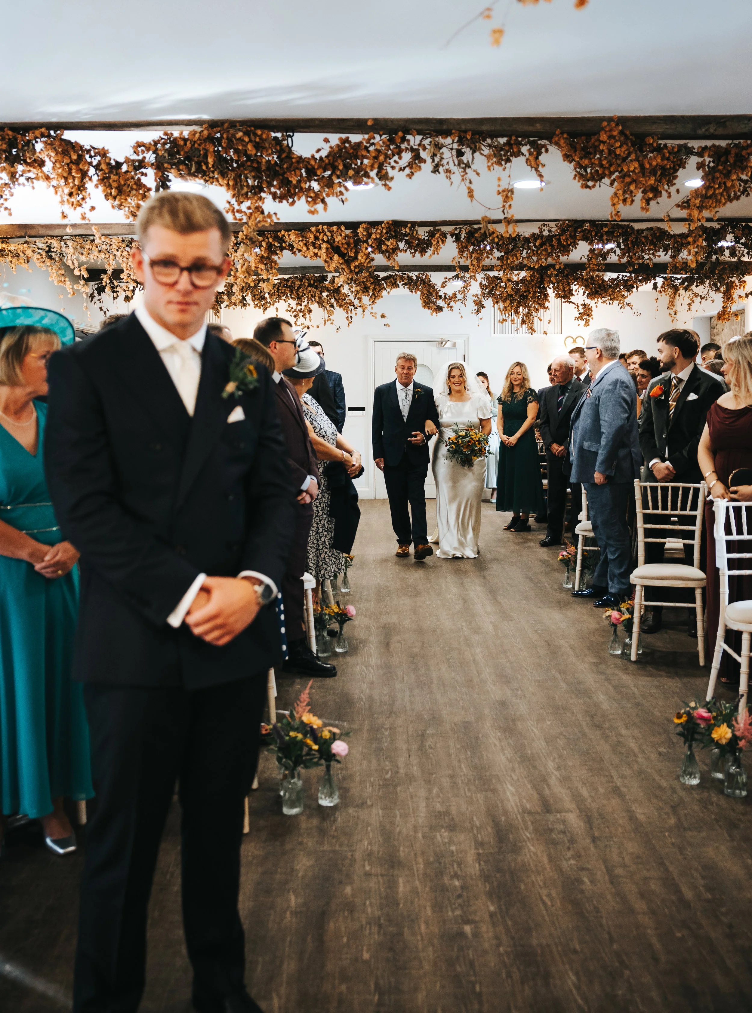 Bride walking down the aisle with her father during a wedding ceremony, surrounded by guests seated on both sides, with floral decorations overhead.