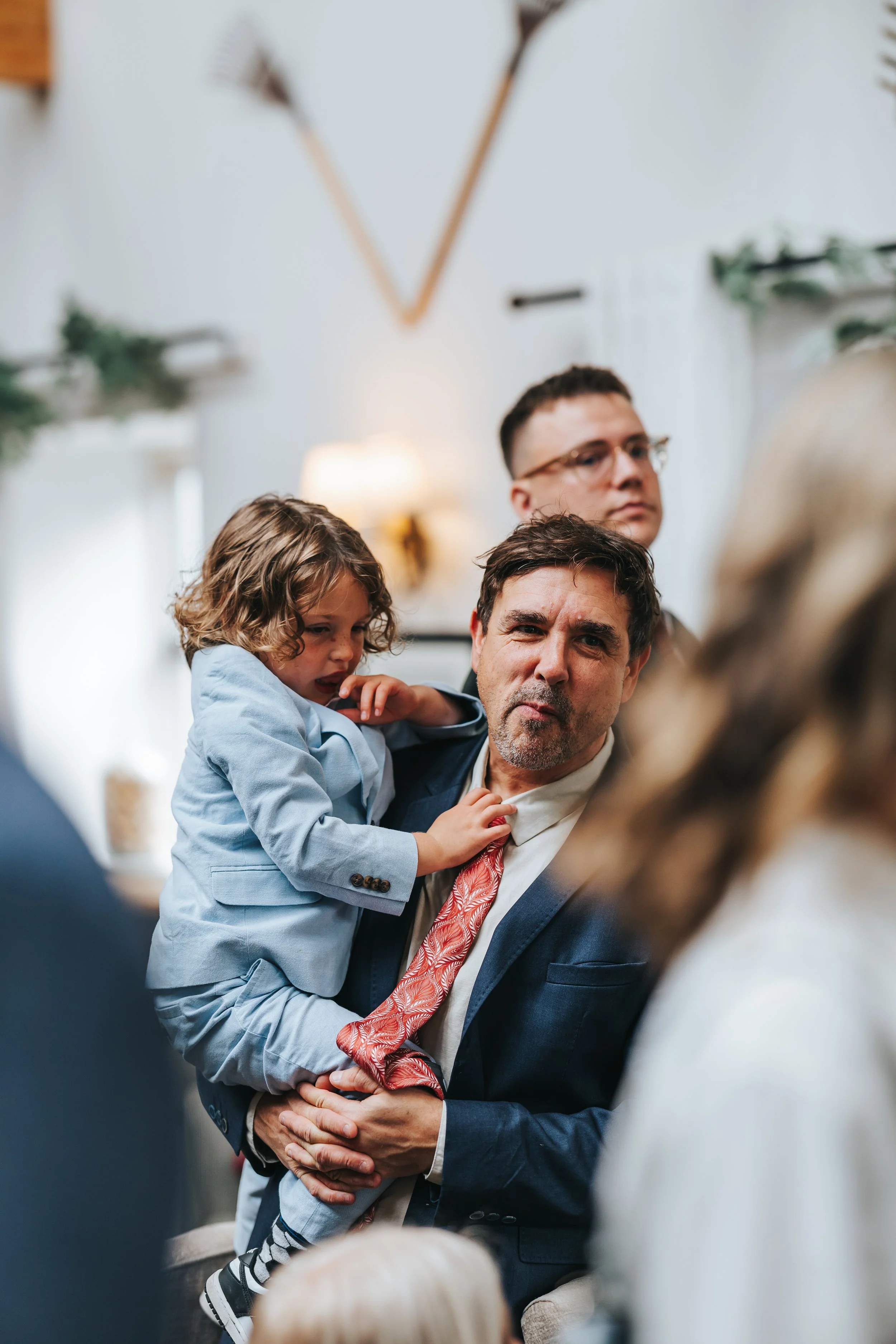 A man in a suit holding a young girl on his lap during a gathering, with a woman in the foreground, a man in glasses in the background, and warm indoor lighting.