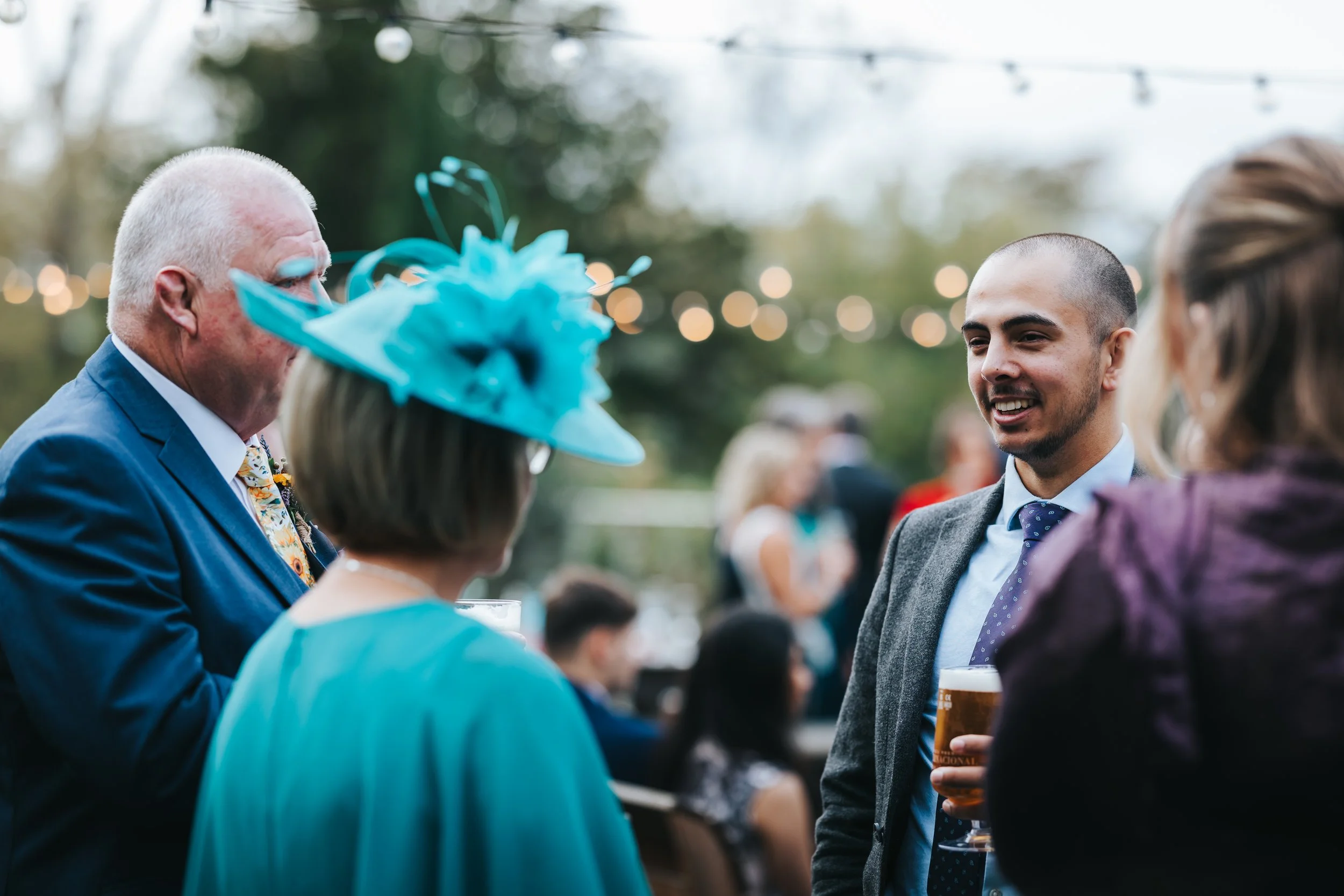 People socializing outdoors at a party, with string lights in the background, including a man in a suit, a woman with a large teal hat, and a man with a drink.