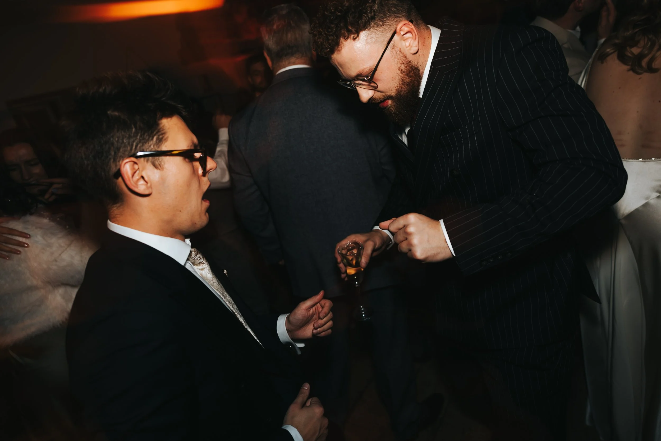 Two men in suits and glasses having a conversation at a social event or party, with other people in the background.