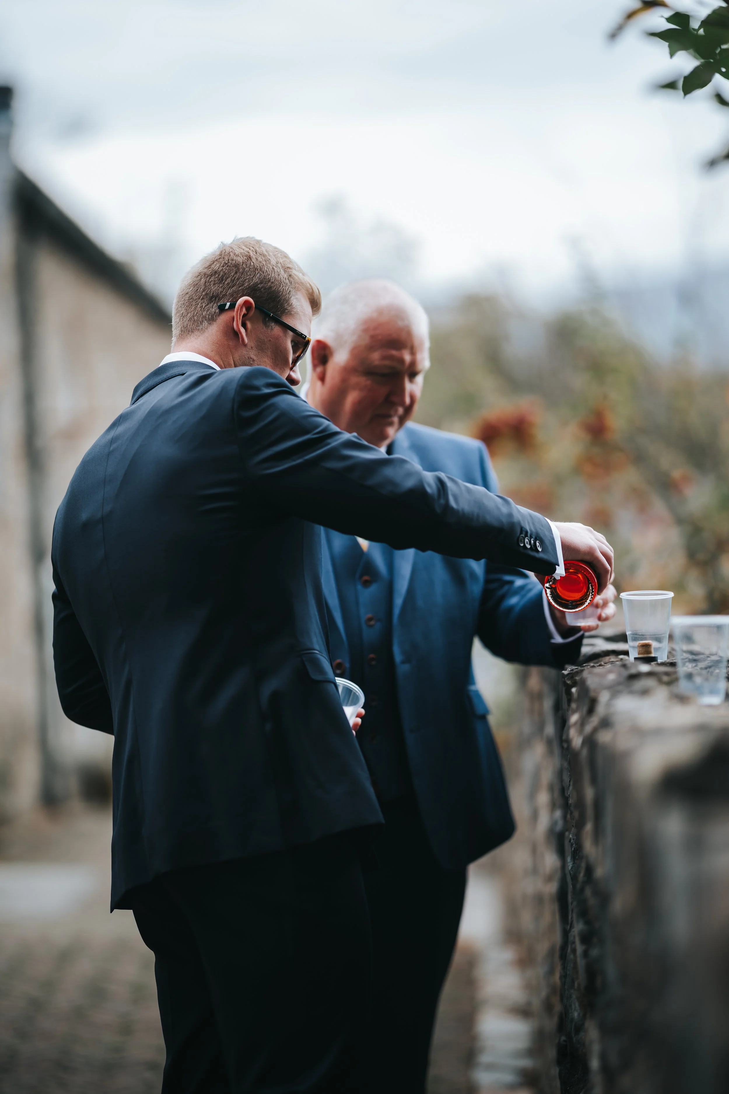 Two men in suits having drinks outdoors against a stone wall with blurry autumn trees in the background.