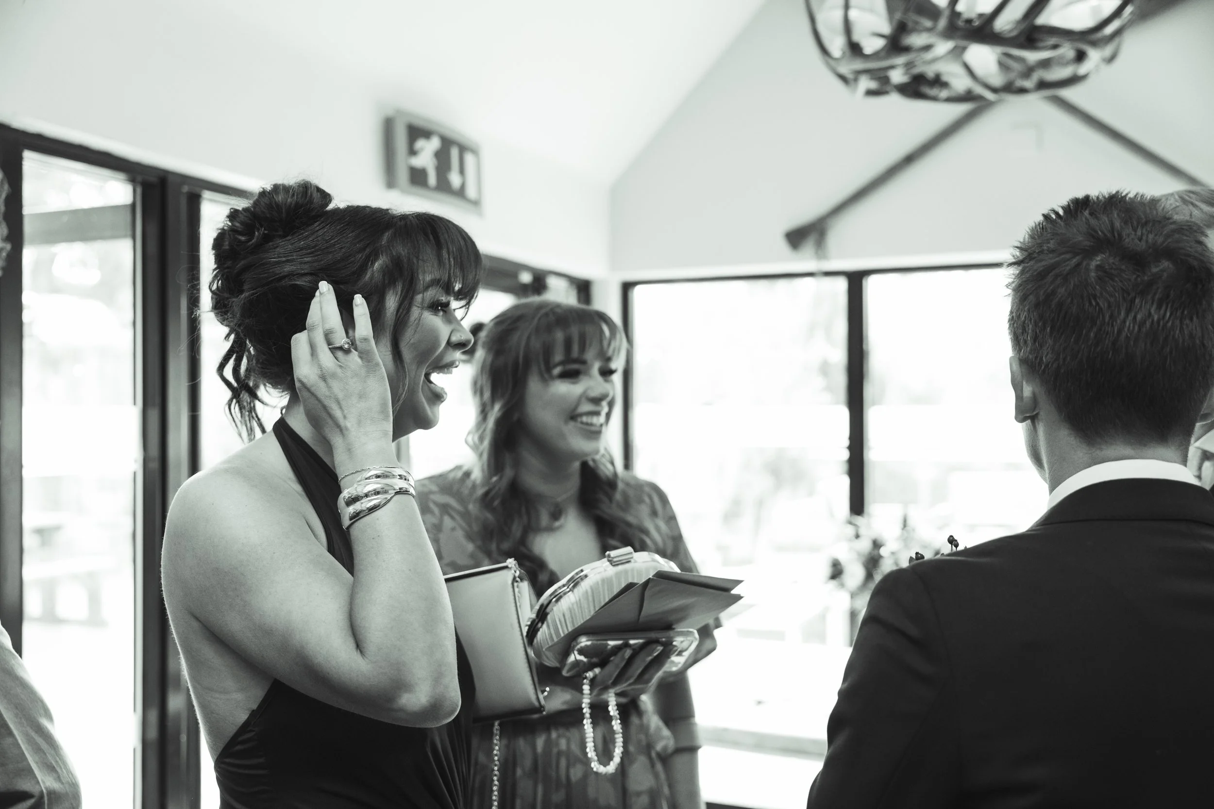Two women and a man engaged in a conversation at an indoor event, with large windows and a sign indicating exit behind them.