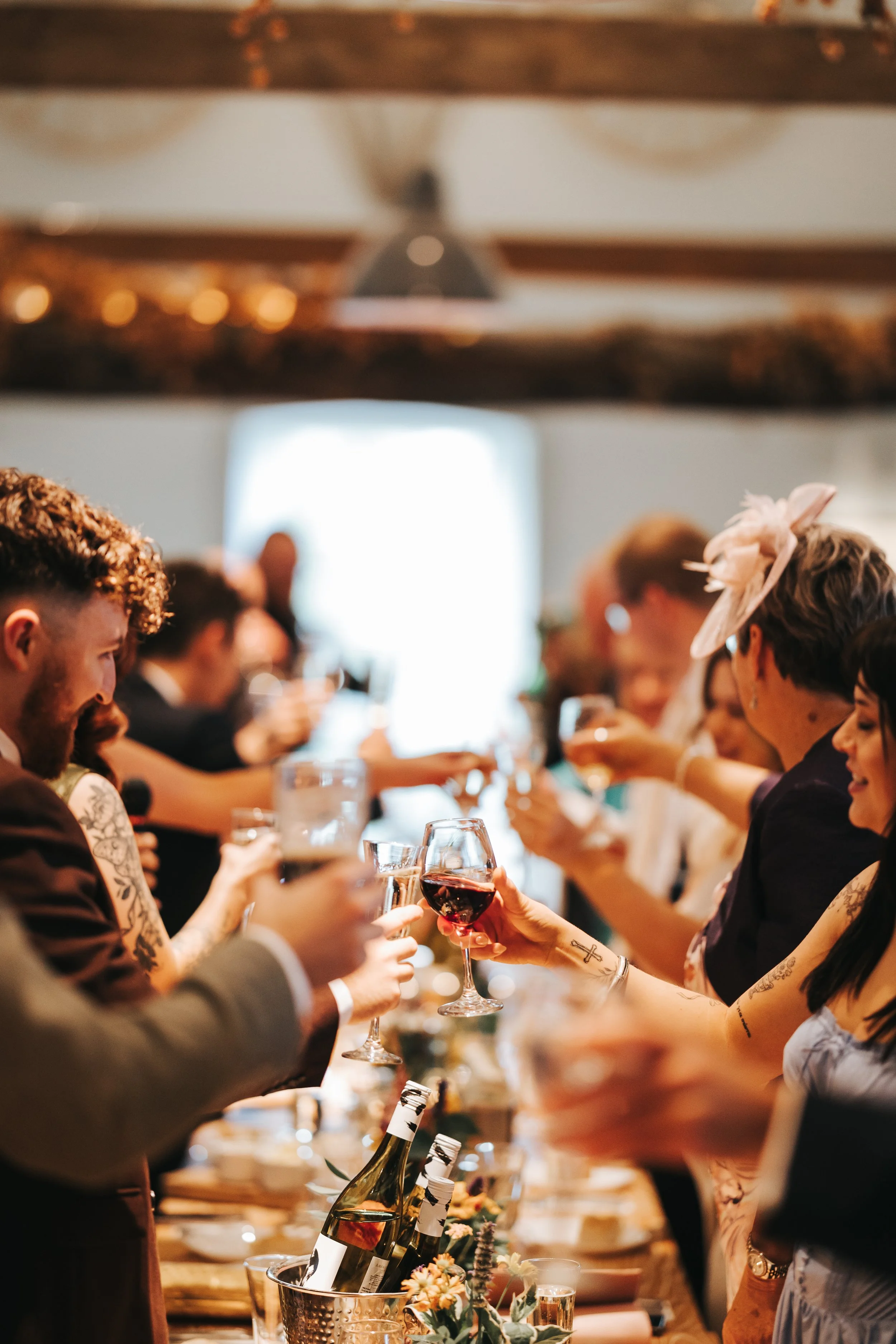 People raising glasses in toast at a celebratory banquet or wedding reception, with bottles of wine and floral centerpieces on the table.