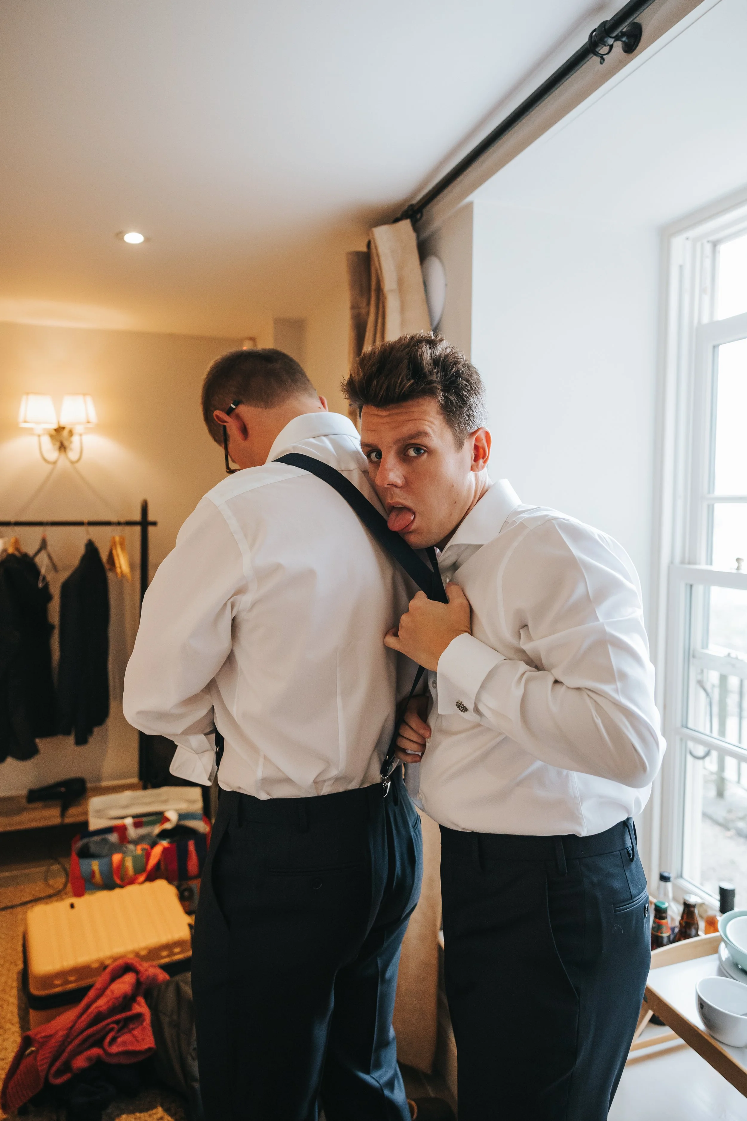 Two men in white shirts and black pants in a room with a window. One man is facing away, the other is holding his suspenders and making a funny face.