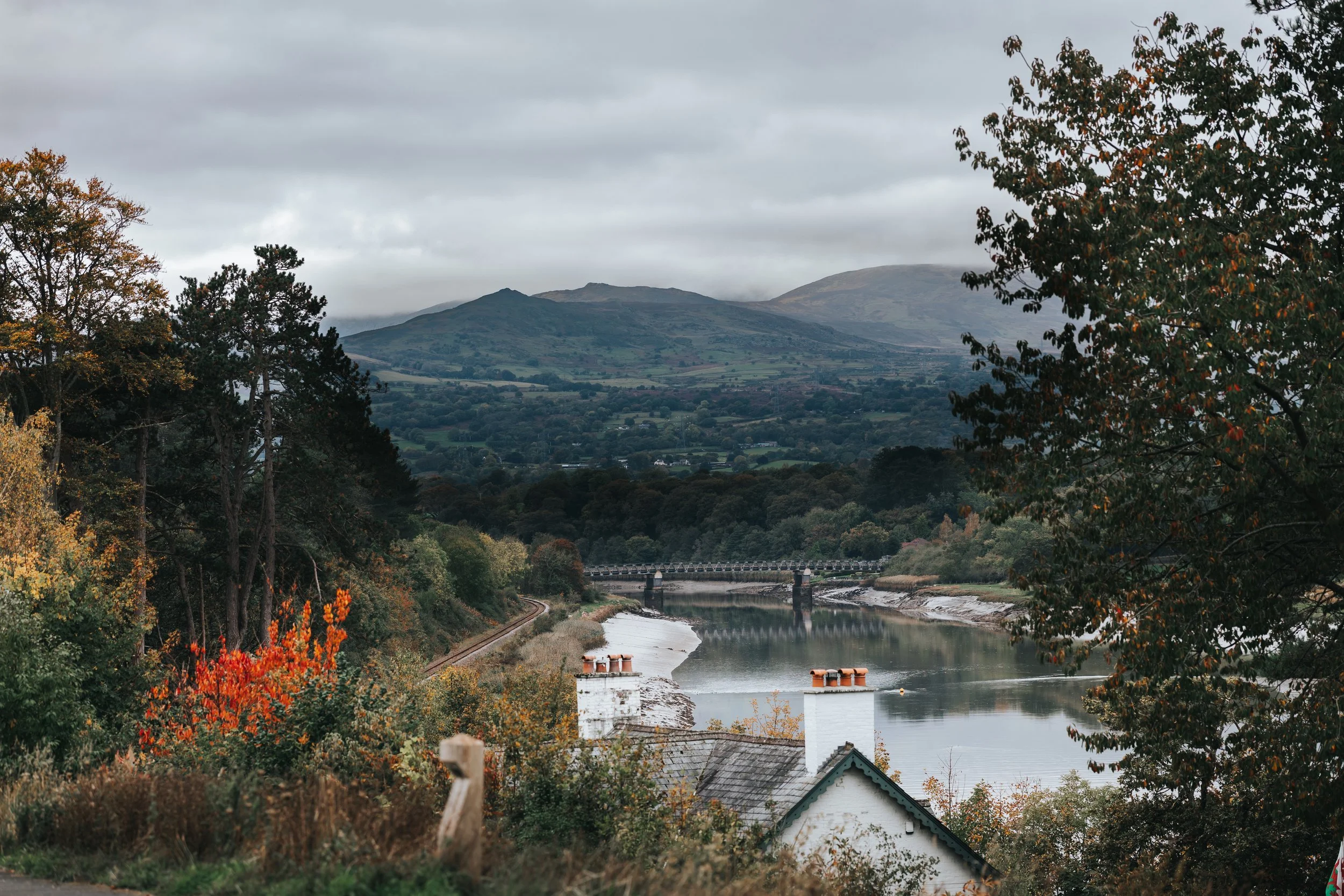 Scenic view of a river with a bridge, surrounded by trees with autumn foliage, mountains in the background, and overcast sky.