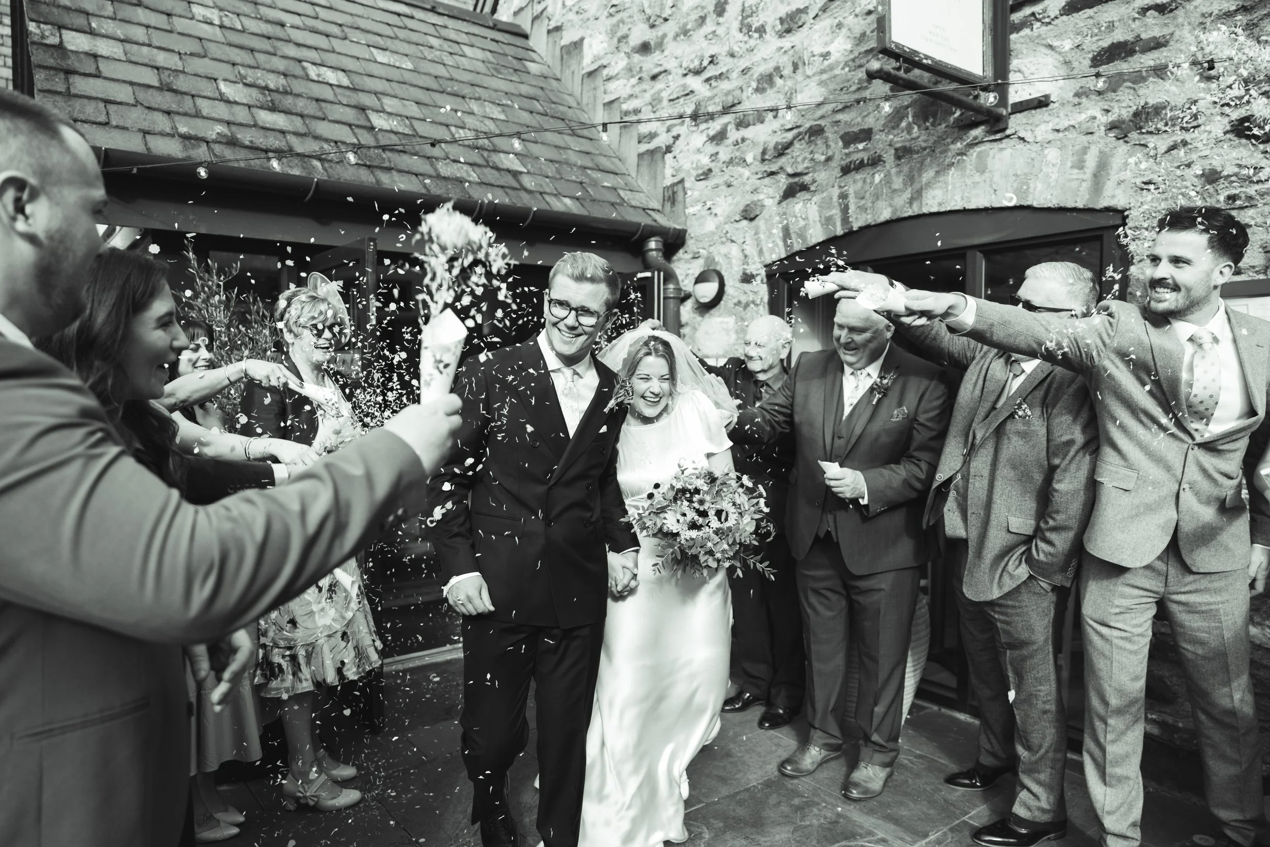 A black and white photo of a wedding celebration with a bride and groom standing hand in hand, surrounded by guests throwing confetti, in a rustic indoor setting.