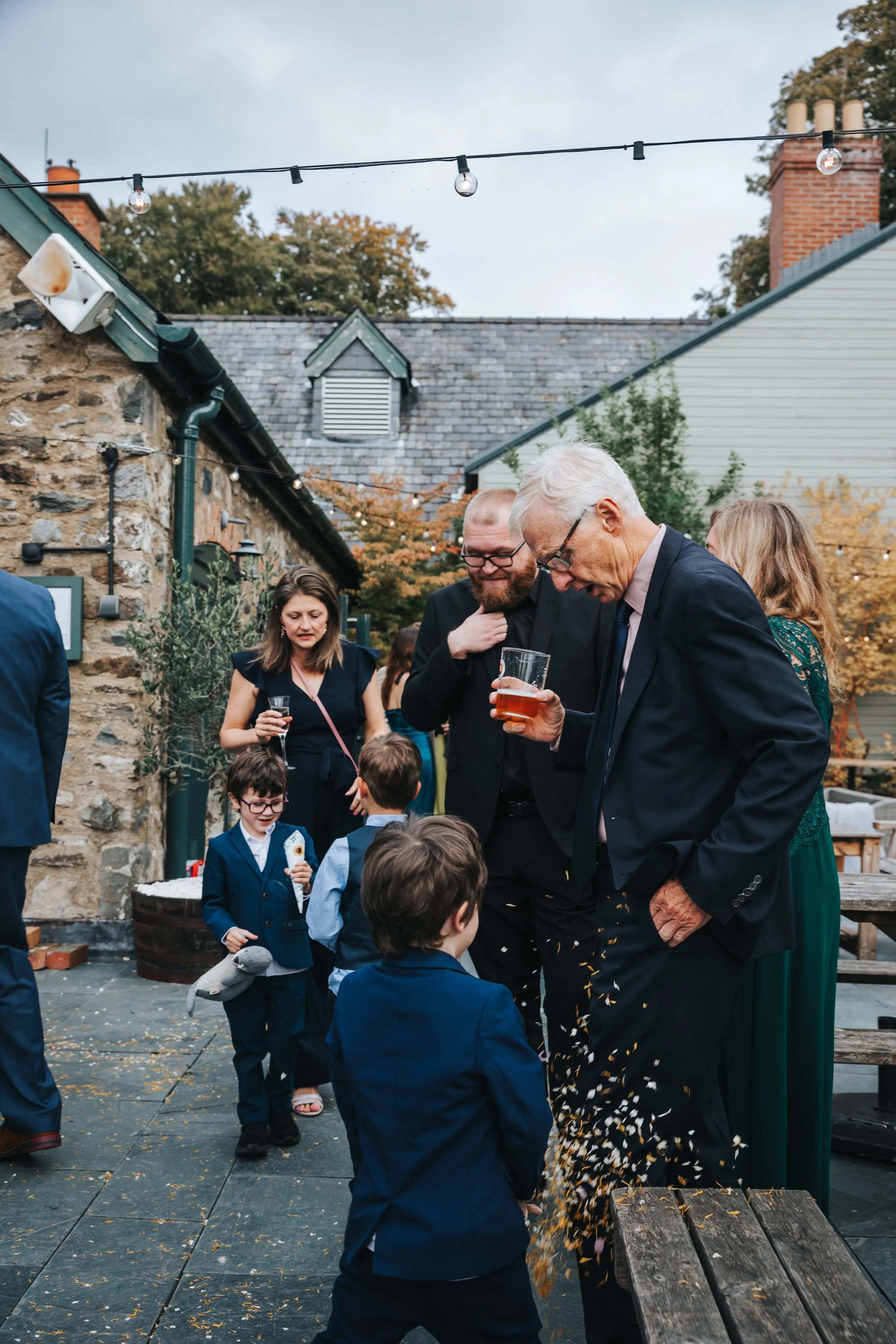 People at a social gathering outdoors, with some children and adults in formal attire, children playing and adults drinking, on a patio decorated with string lights and surrounded by stone and wooden buildings, during early evening.