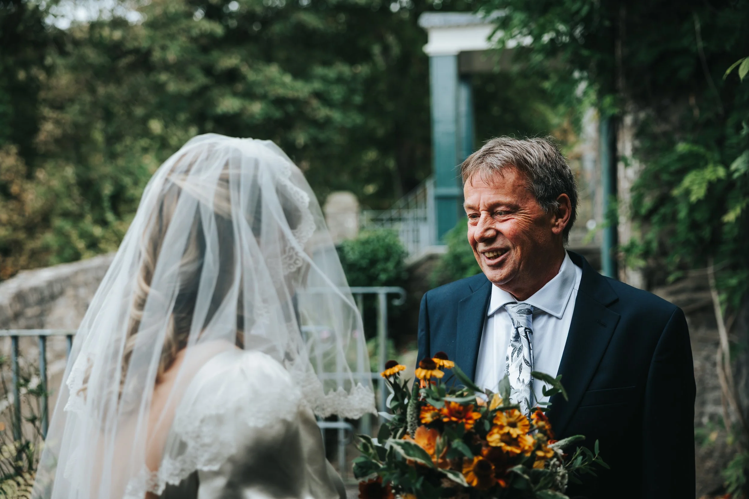A bride in a white wedding dress and veil holding a bouquet of flowers, smiling and talking to an older man in a suit outside in a green, wooded area.