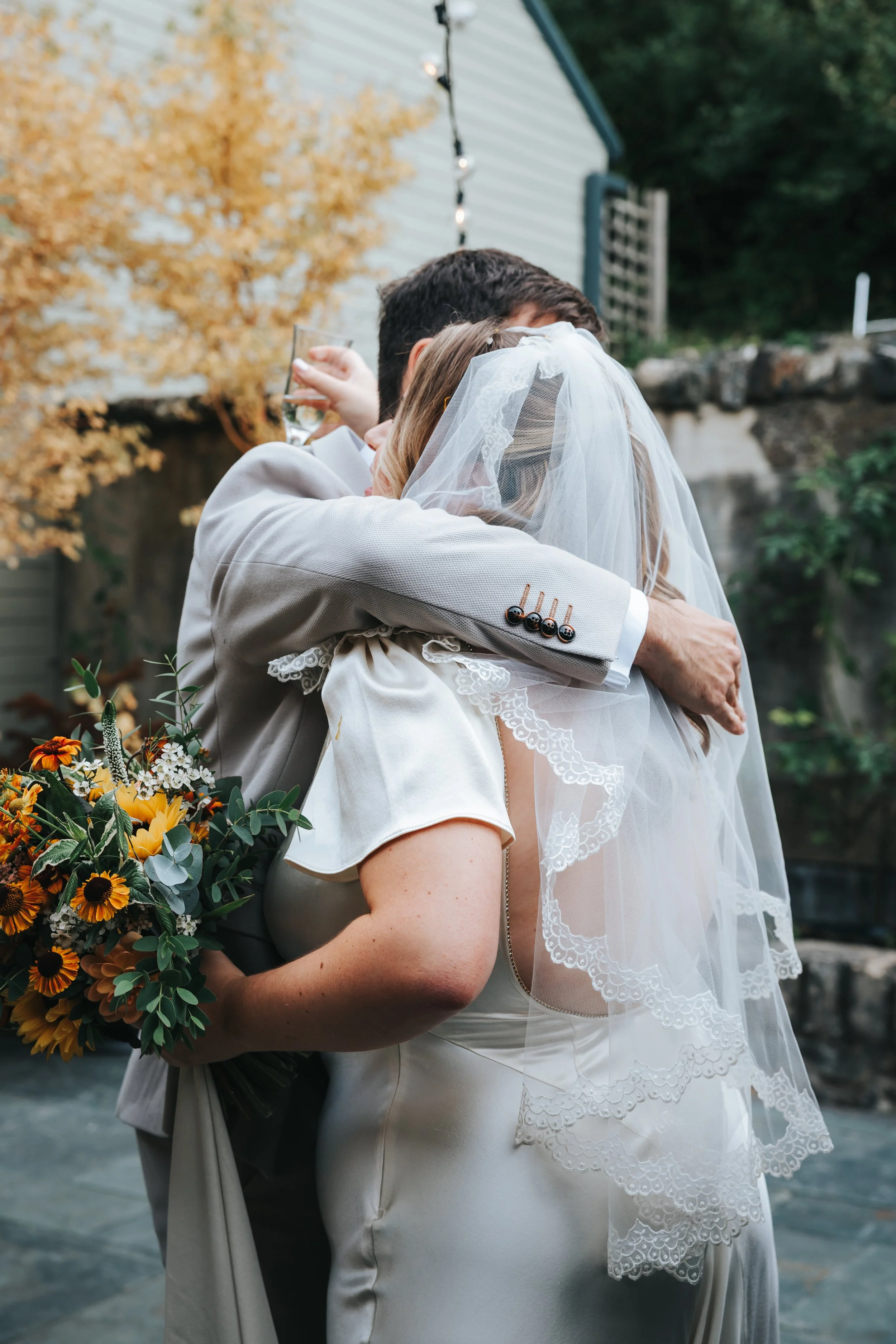 A bride and groom hugging outdoors at a wedding, with the bride holding a colorful bouquet and wearing a veil, the groom in a light-colored suit, and a tree with autumn leaves in the background.