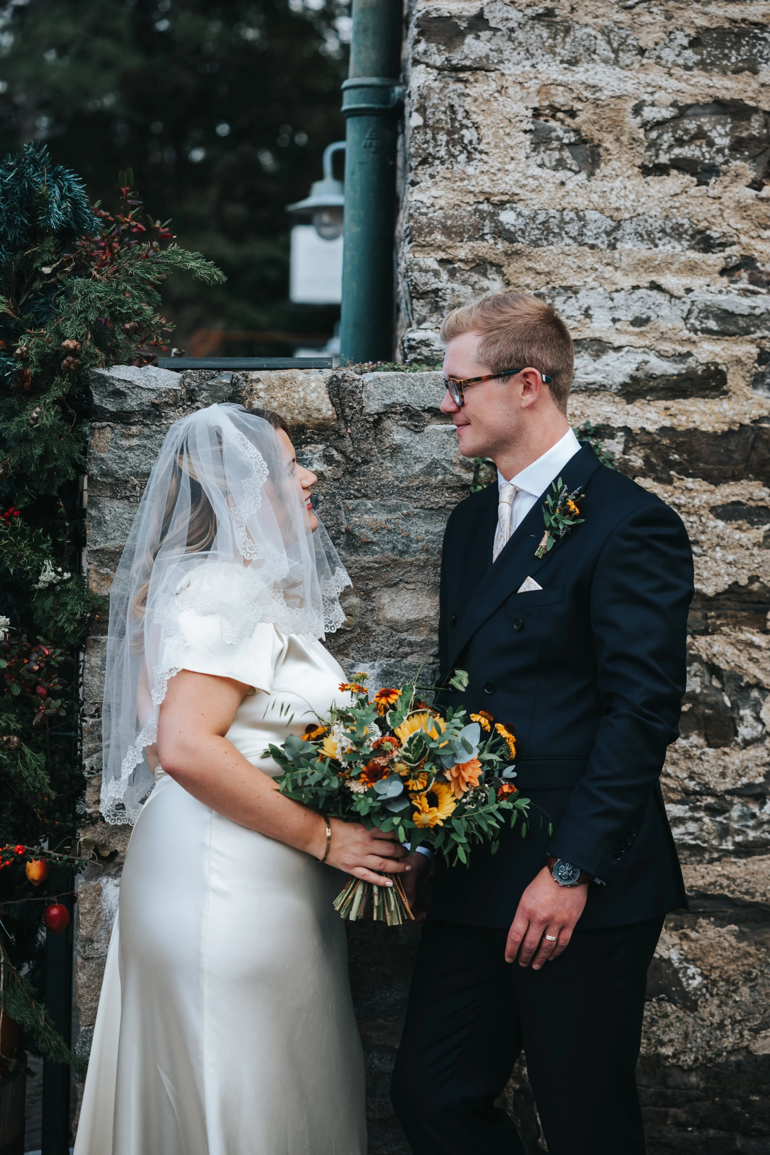 A bride and groom standing close together outdoors near a stone wall, smiling at each other. The bride is wearing a white satin dress with a lace-edged veil and holding a bouquet of yellow, orange, and green flowers. The groom is wearing a dark suit 