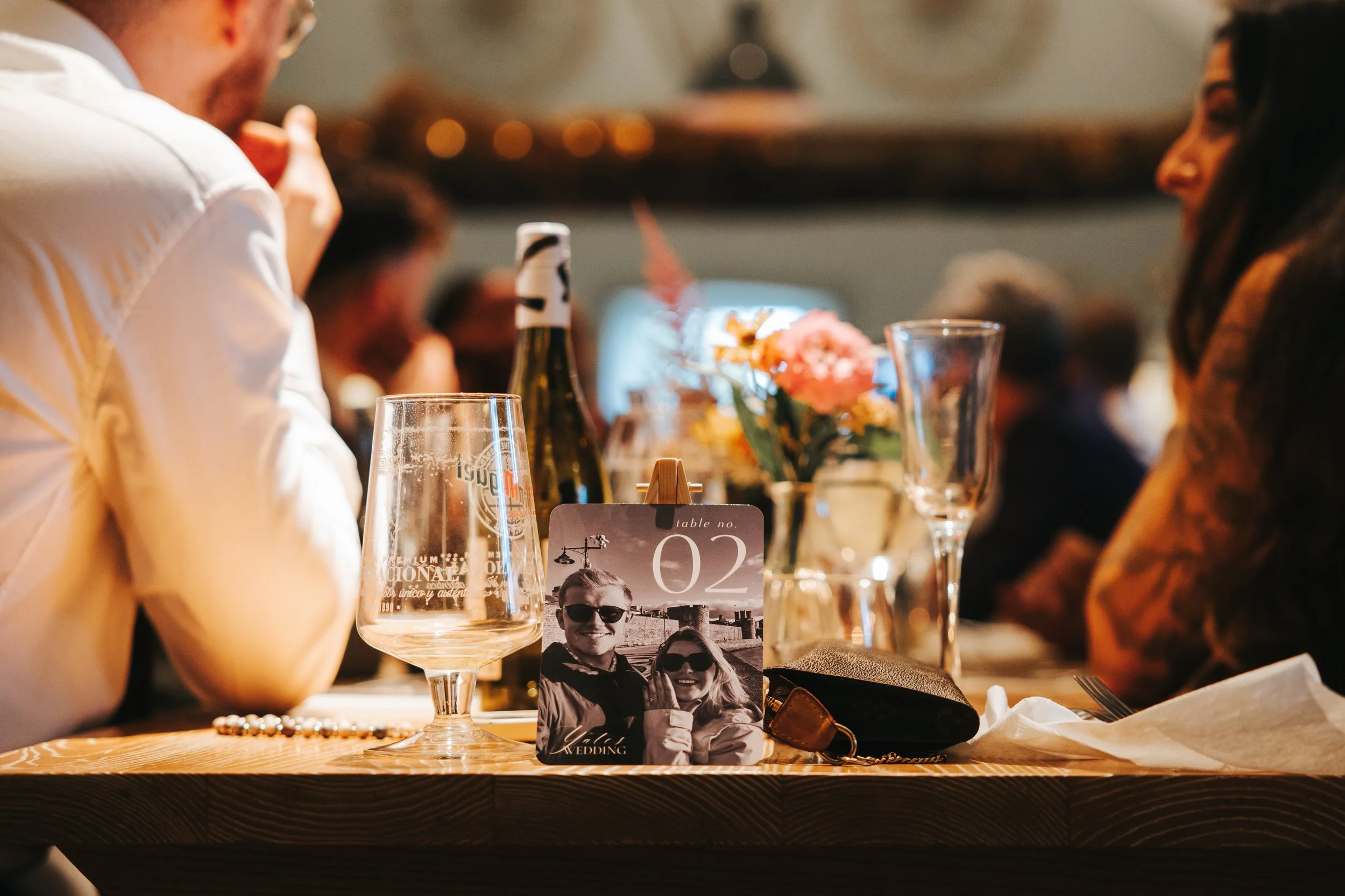 A table at a wedding reception with a photo booth sign, empty wine glasses, a bouquet of flowers, and a purse. People are sitting and talking in the background.