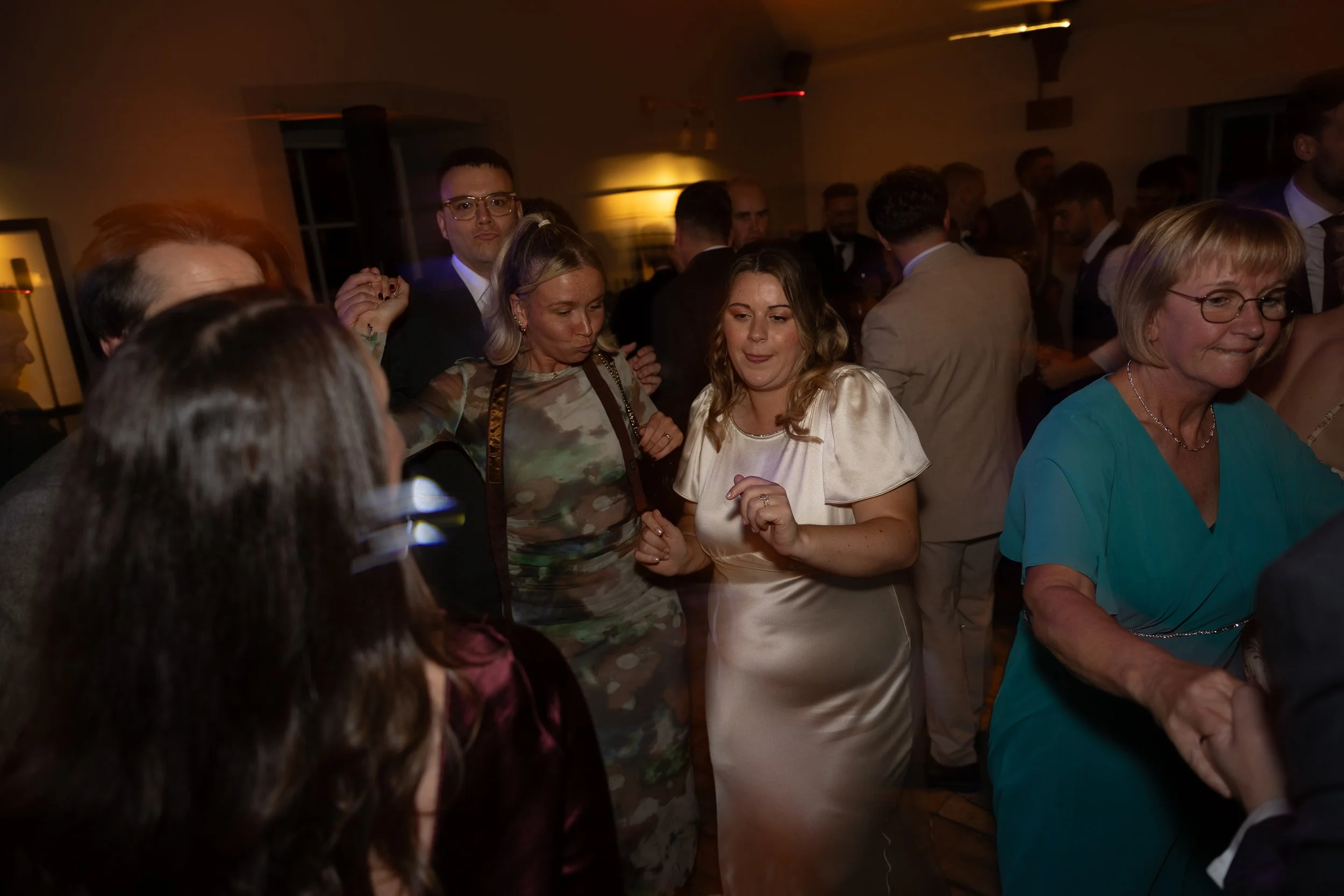 People dancing at a party, with a woman in a shiny cream dress in the center, surrounded by others, in a dimly lit room.