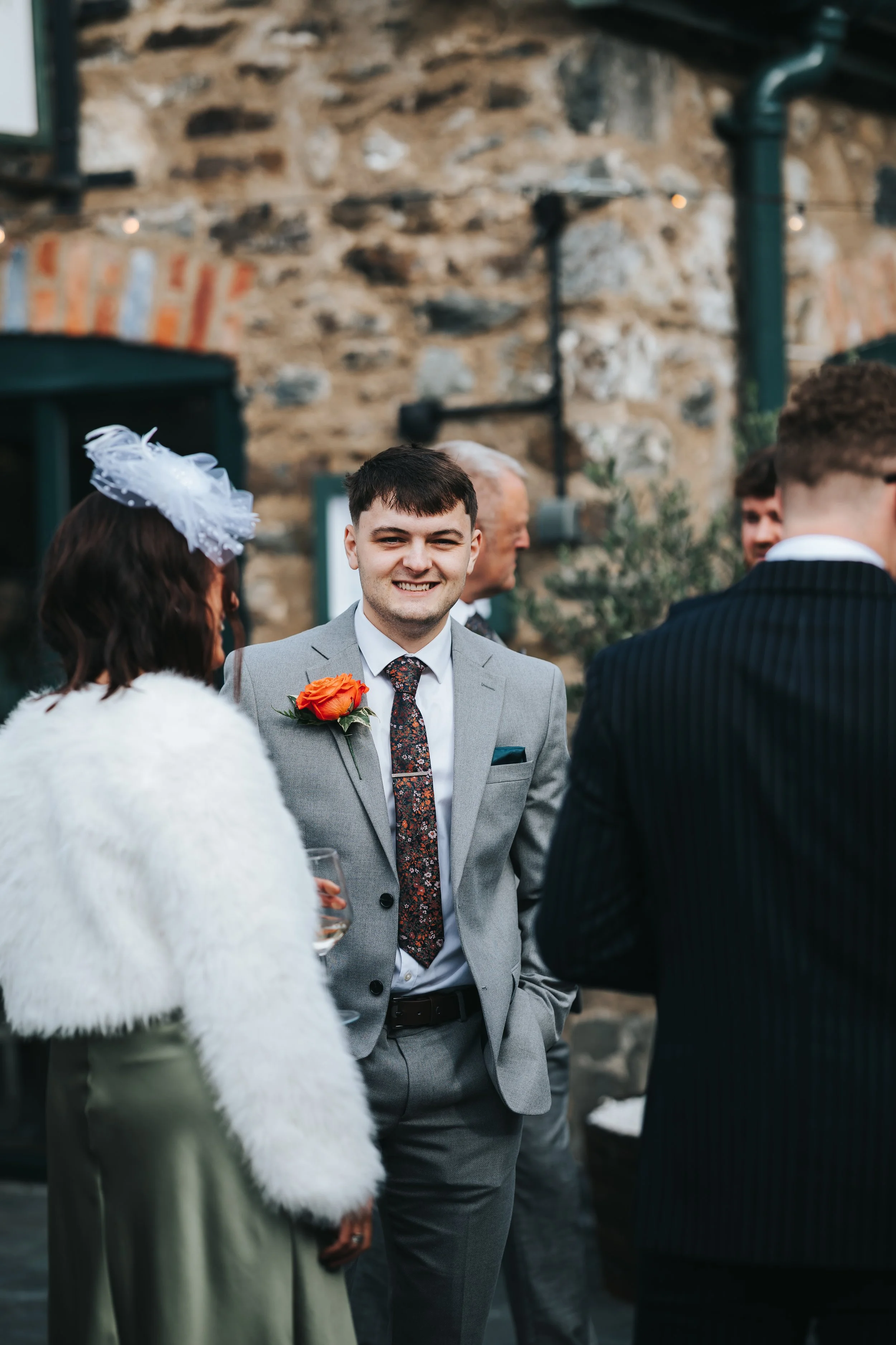 A smiling man in a gray suit with a floral tie and a orange flower pinned to his lapel, standing outdoors with people dressed in formal attire and a stone building in the background.
