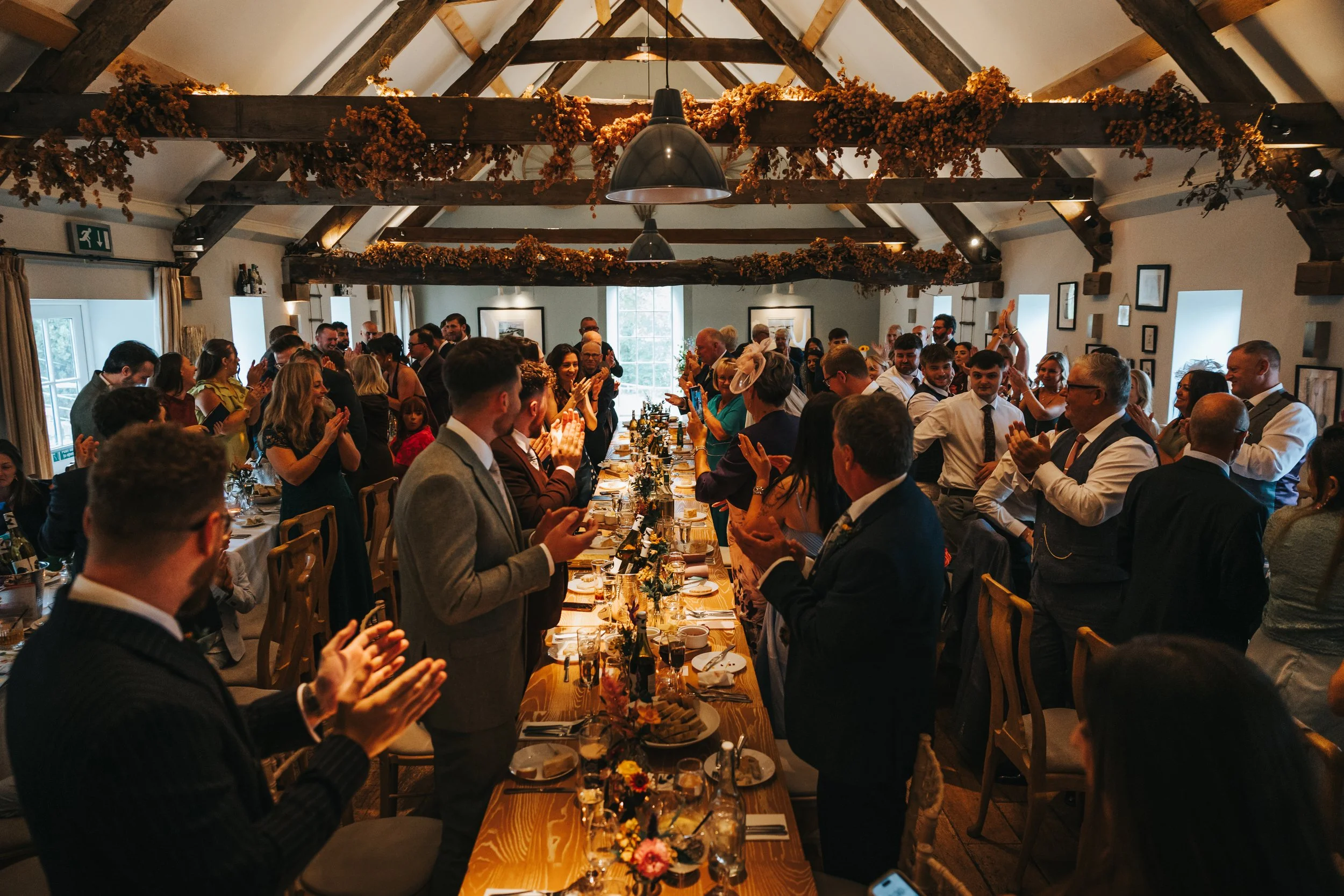 A large group of people gathered in a rustic banquet hall celebrating, standing around a long decorated table with food and drinks, clapping and enjoying the event.