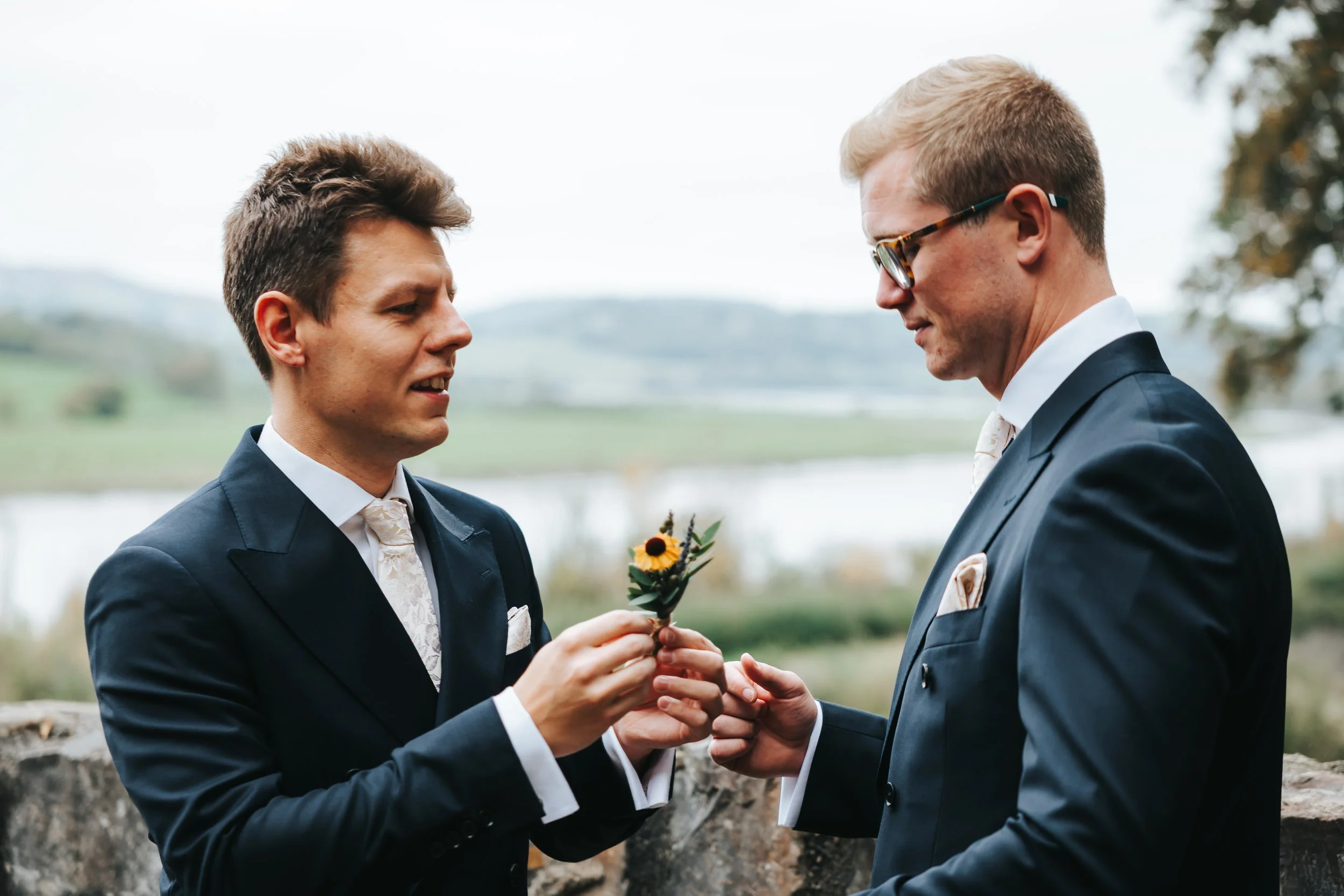 Two men in suits exchanging a boutonniere outdoors by a stone wall, with water and hillside in the background.
