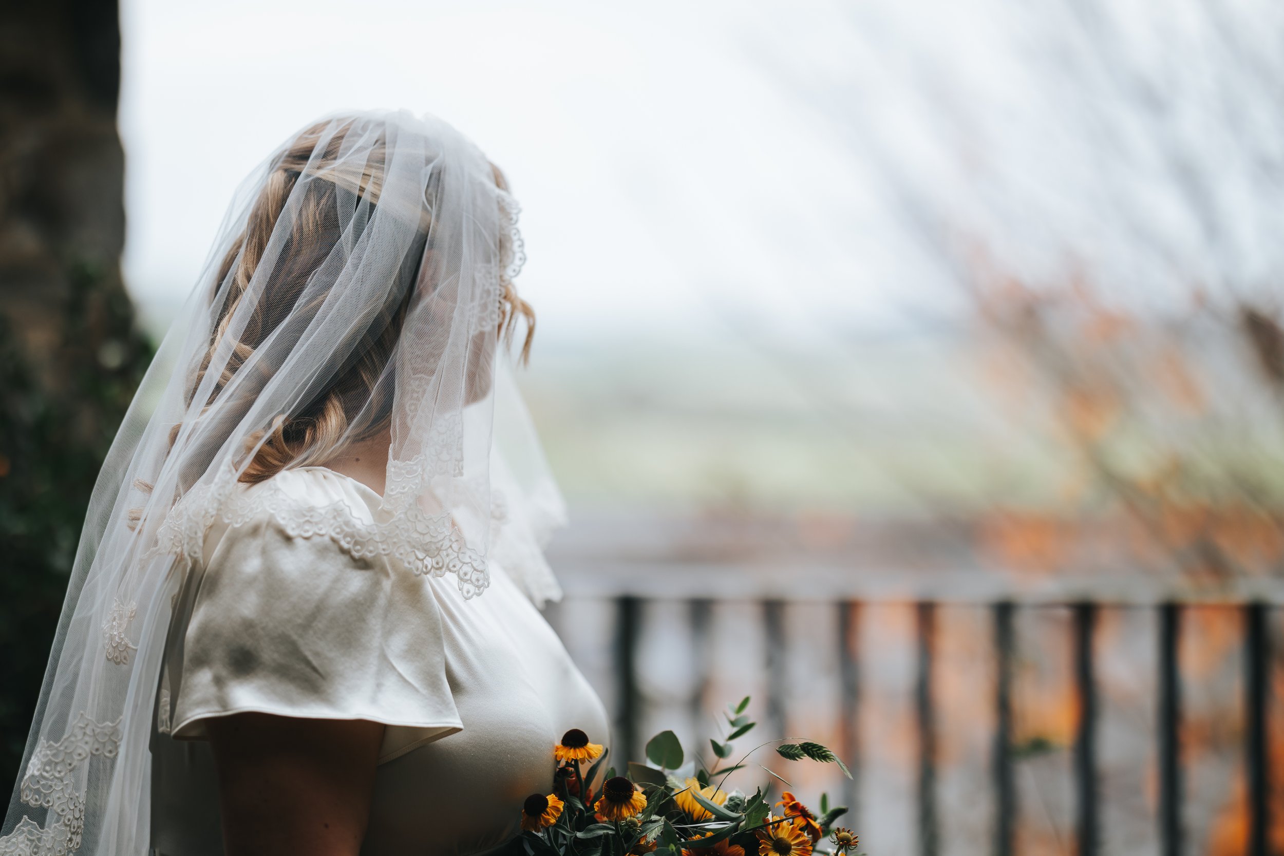 A woman in a wedding dress and veil holding a bouquet of yellow and dark flowers, standing on a balcony with blurred fall foliage in the background.