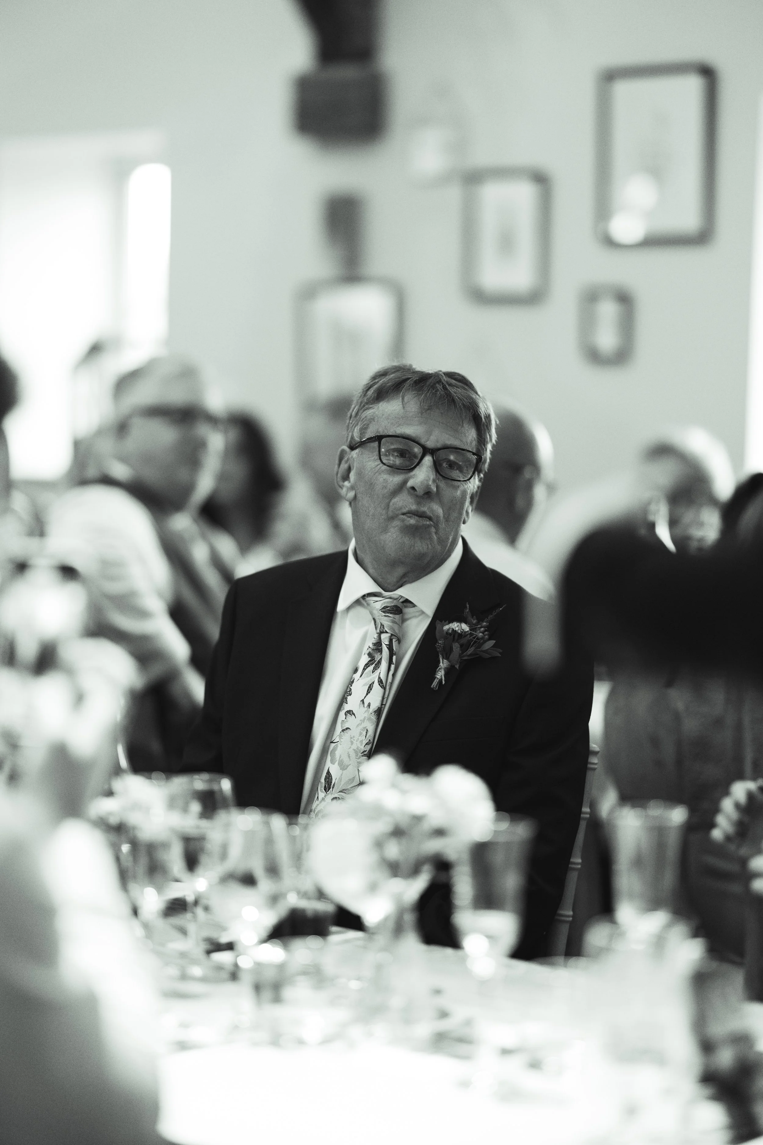 A black and white photo of an older man with glasses, wearing a suit with a floral tie and boutonnière, seated at a banquet table with blurred people and framed pictures on the wall in the background.