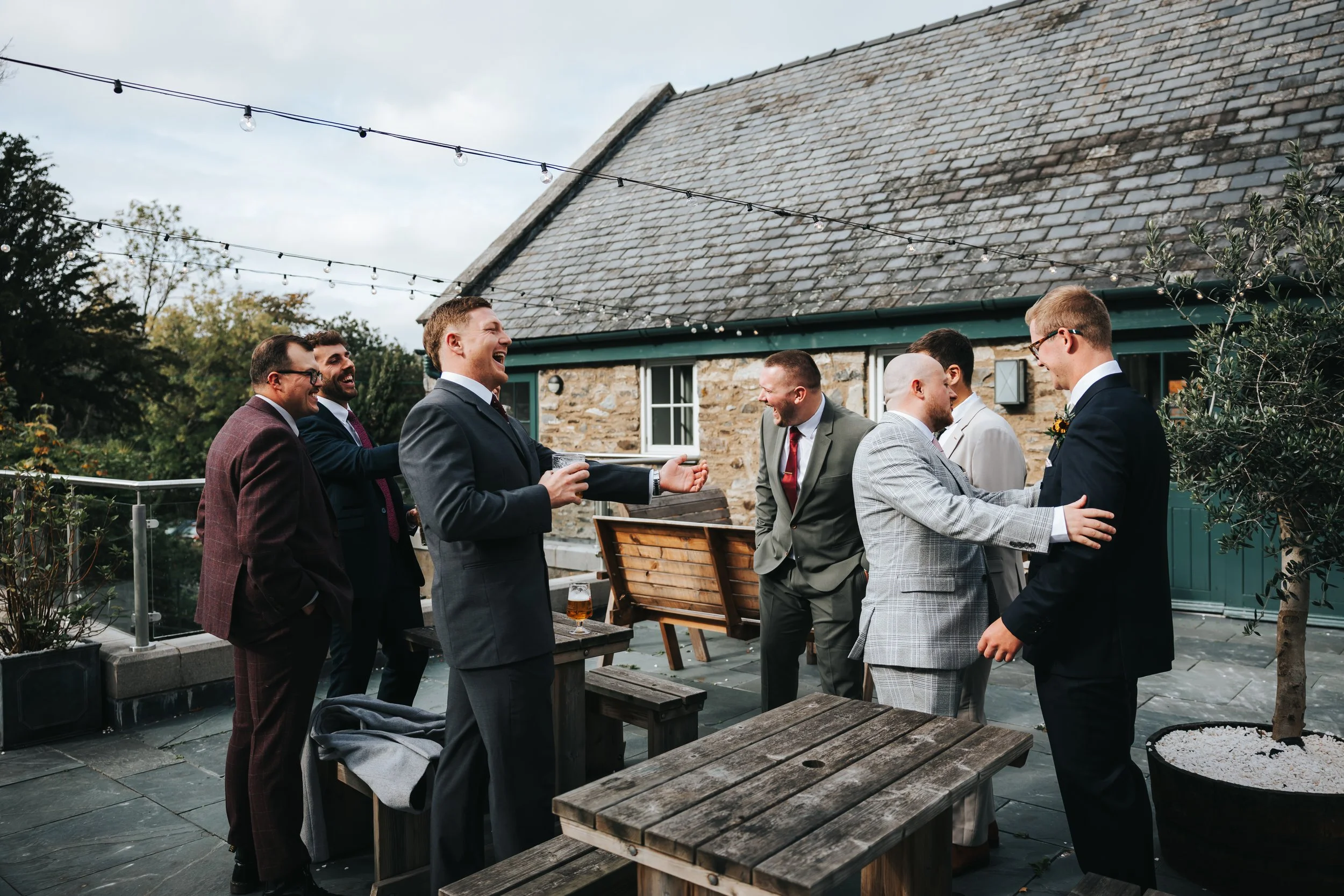 Group of men in suits socializing and laughing on a rooftop patio with string lights and rustic stone building in the background.