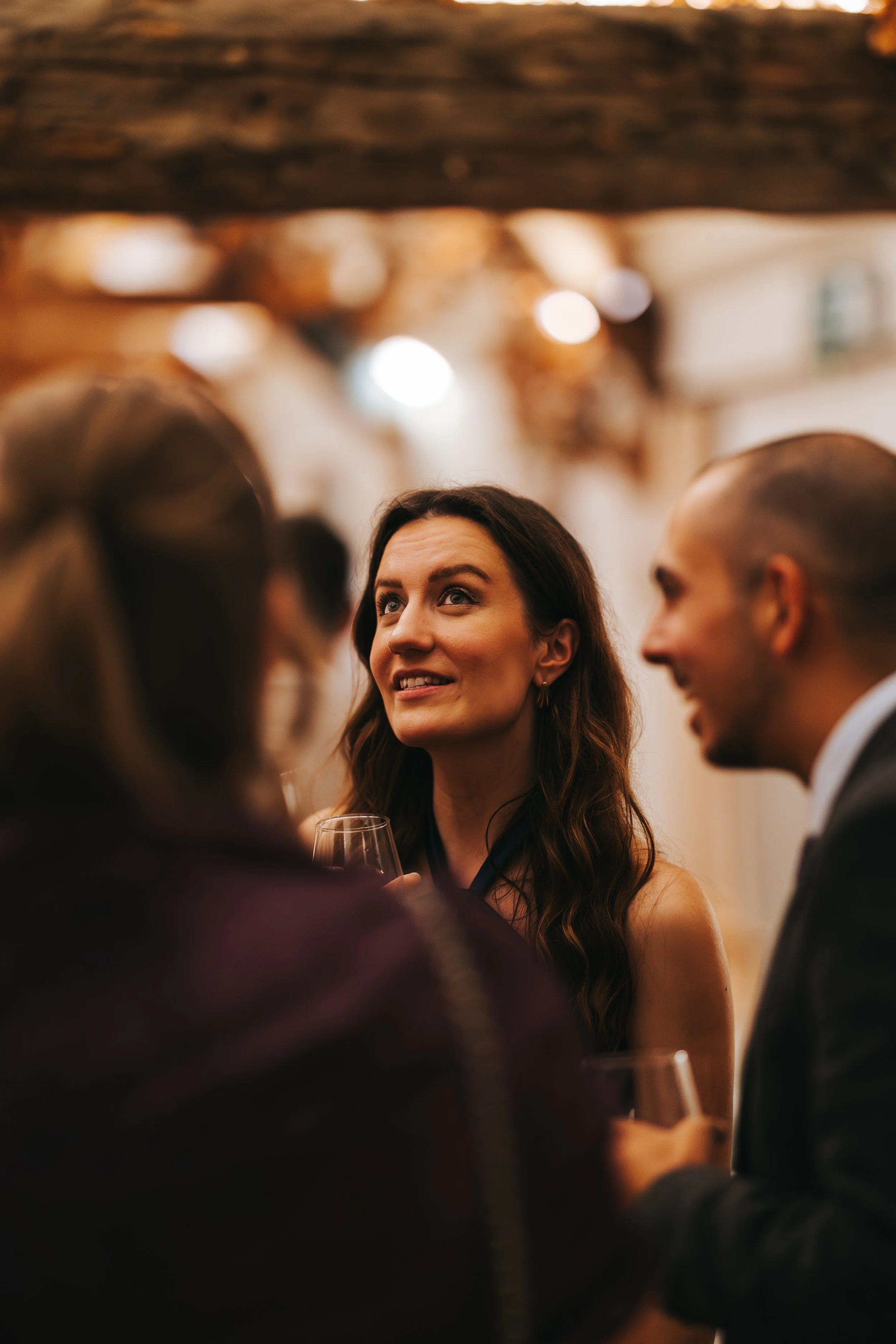 A woman holding a wine glass, speaking with a man and two others at a social gathering or party in an indoor setting with warm lighting.