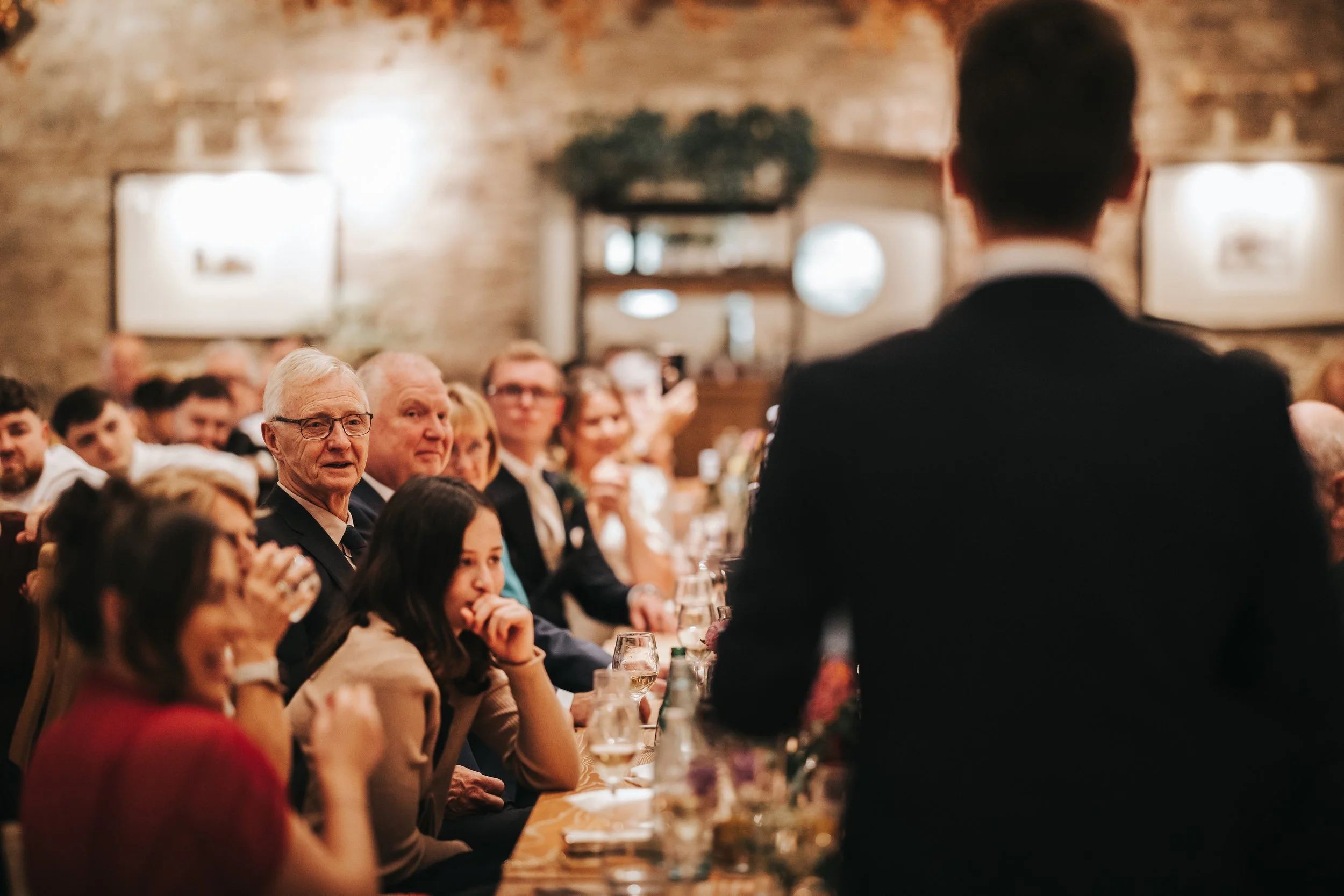 A man giving a speech to an audience in a restaurant or event space with exposed brick walls and artwork, with guests seated at a long table listening attentively.