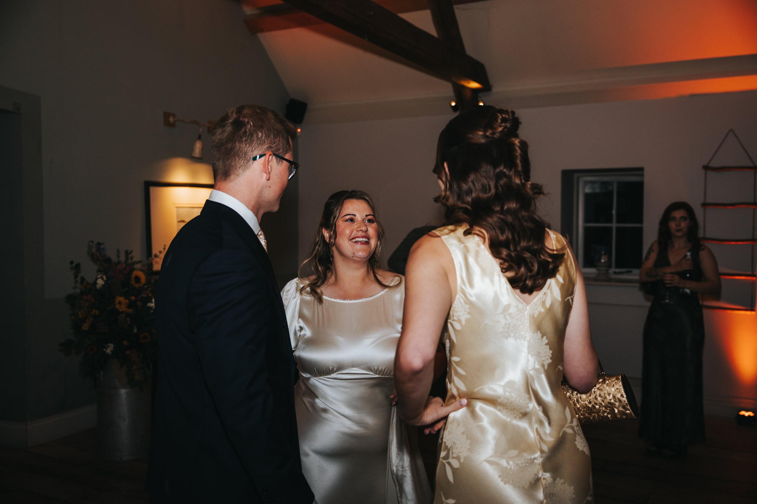 People at a social event, chatting and smiling indoors with warm lighting, floral arrangement in the background, and a woman standing by a window in the background.