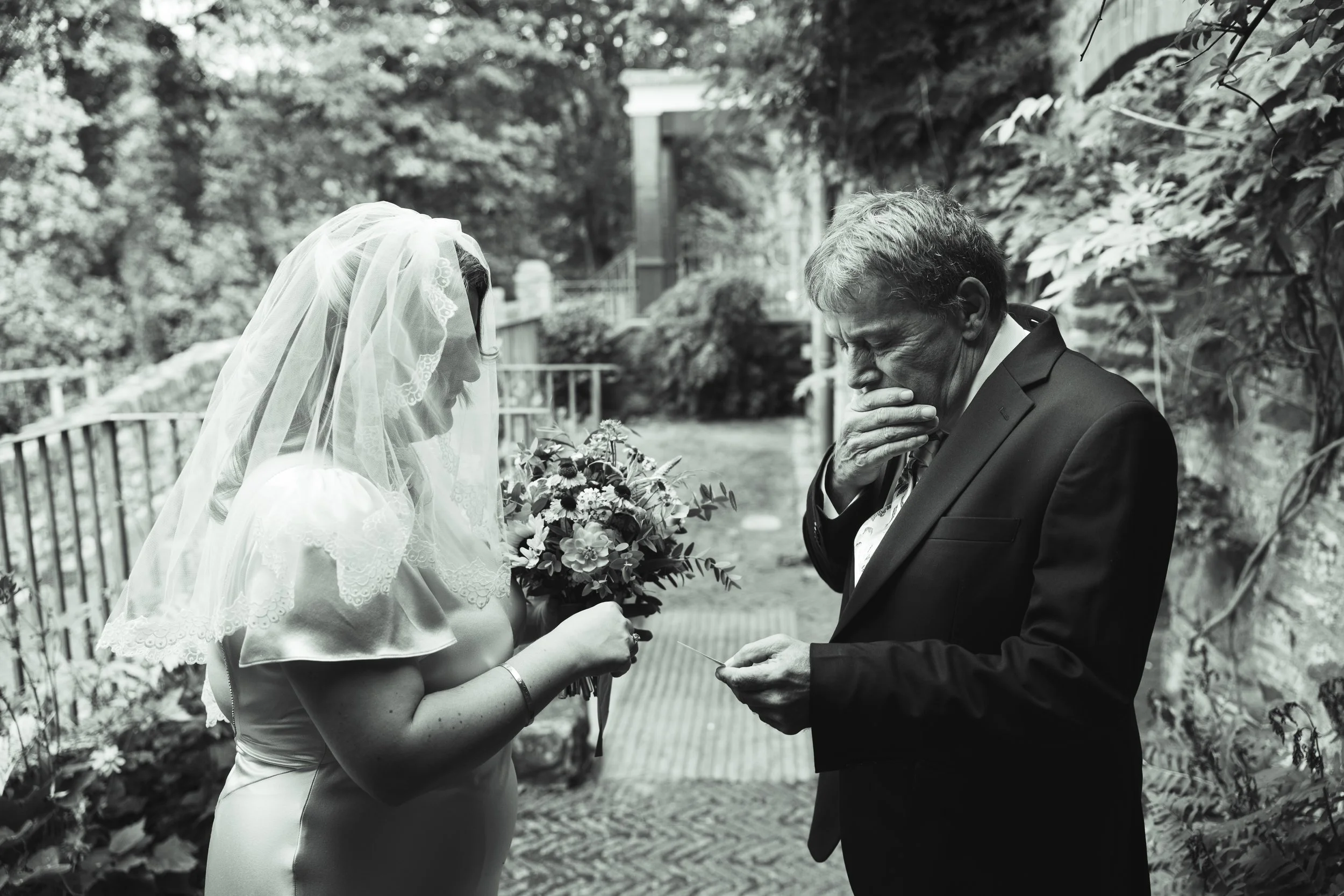 A bride and groom during a wedding ceremony outdoors. The bride is wearing a wedding dress and veil, holding a bouquet of flowers, and the groom is in a suit, holding a ring and showing an emotional reaction.