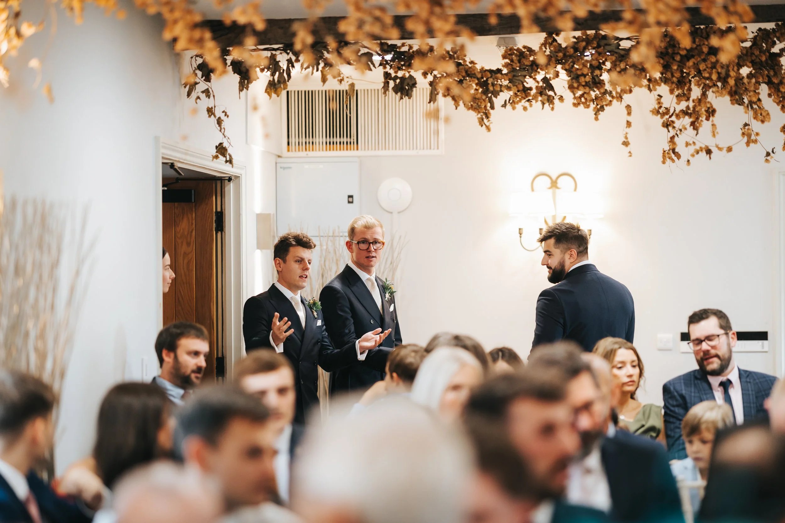 A wedding reception room with beige walls, decorated with hanging dried flowers or leaves. Several guests are seated, and three men in suits are standing and speaking near a doorway. One man is facing away, while the others are looking toward the aud