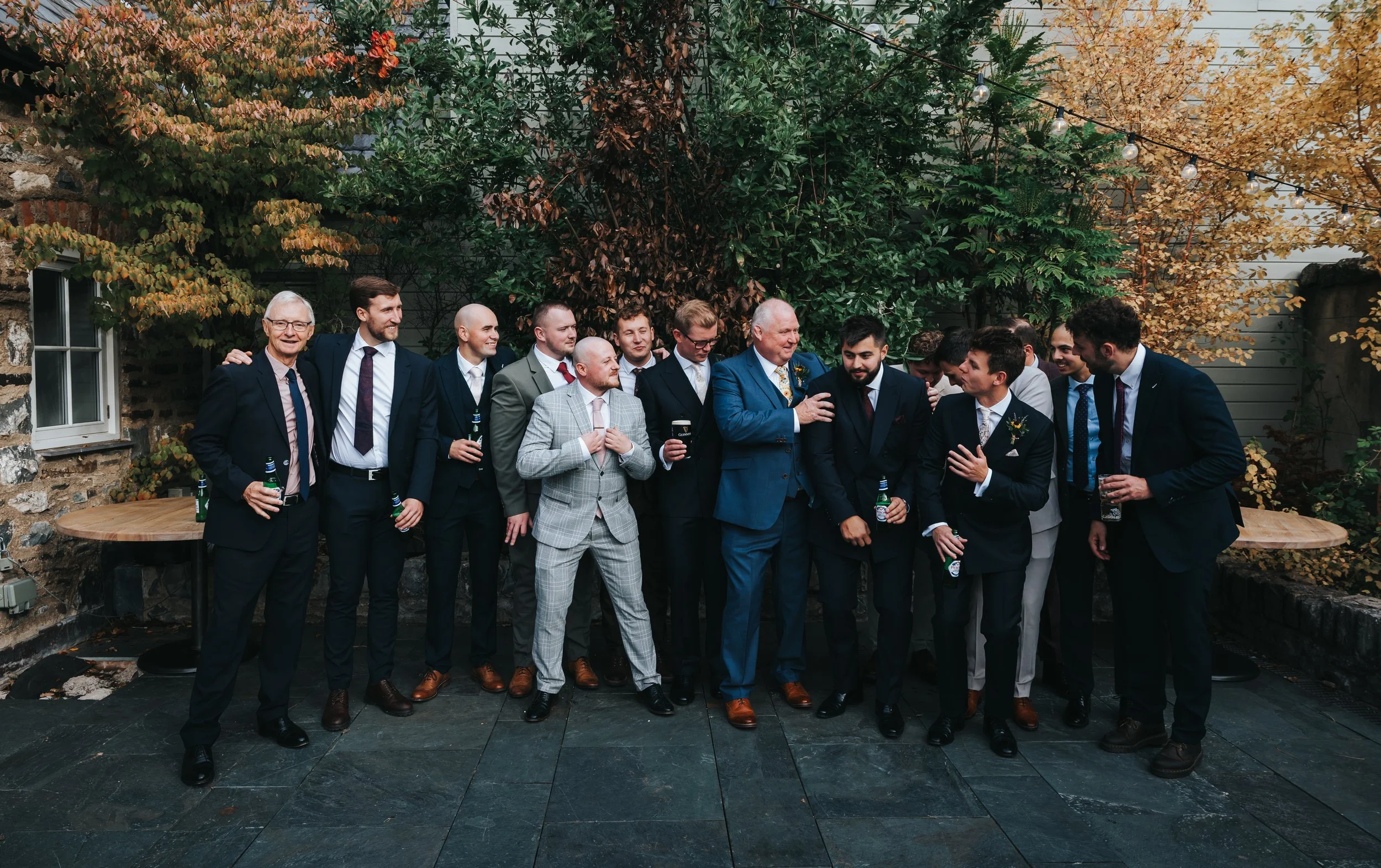Group of men in suits at an outdoor gathering, some holding drinks, engaged in conversation. Autumn trees and string lights in the background.