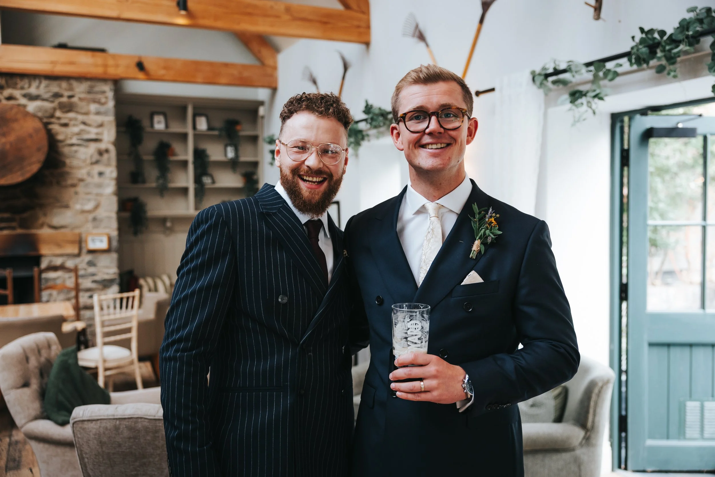 Two men dressed in formal suits smiling indoors, one holding a glass of beer. The background features a stone wall, wooden beams, and a doorway with white curtains, decorated with greenery.