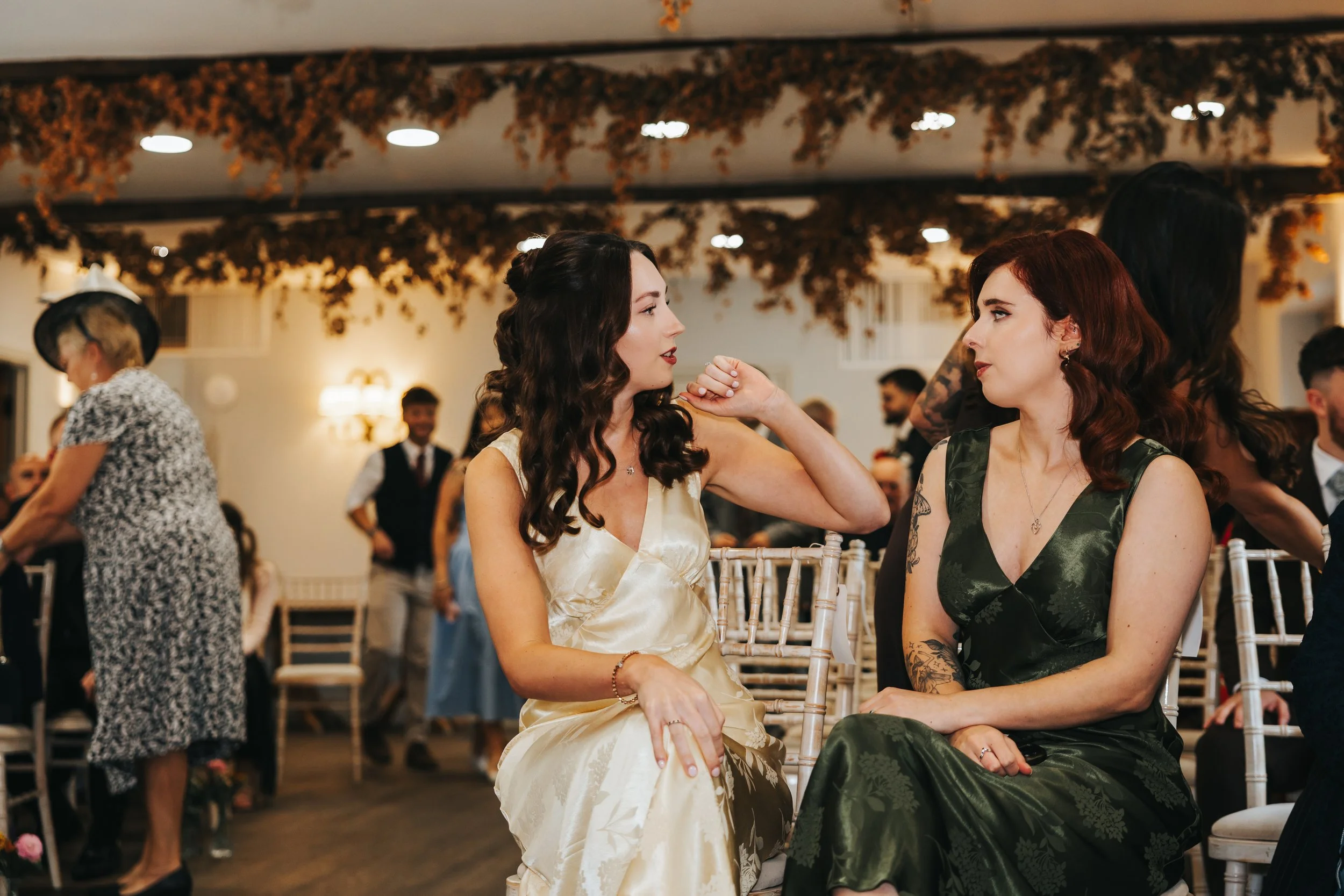 Two women sitting on chairs at a wedding or formal event, engaging in a conversation. The woman on the left has long curly brown hair and is wearing a cream satin dress, while the woman on the right has shoulder-length red hair and is wearing a green