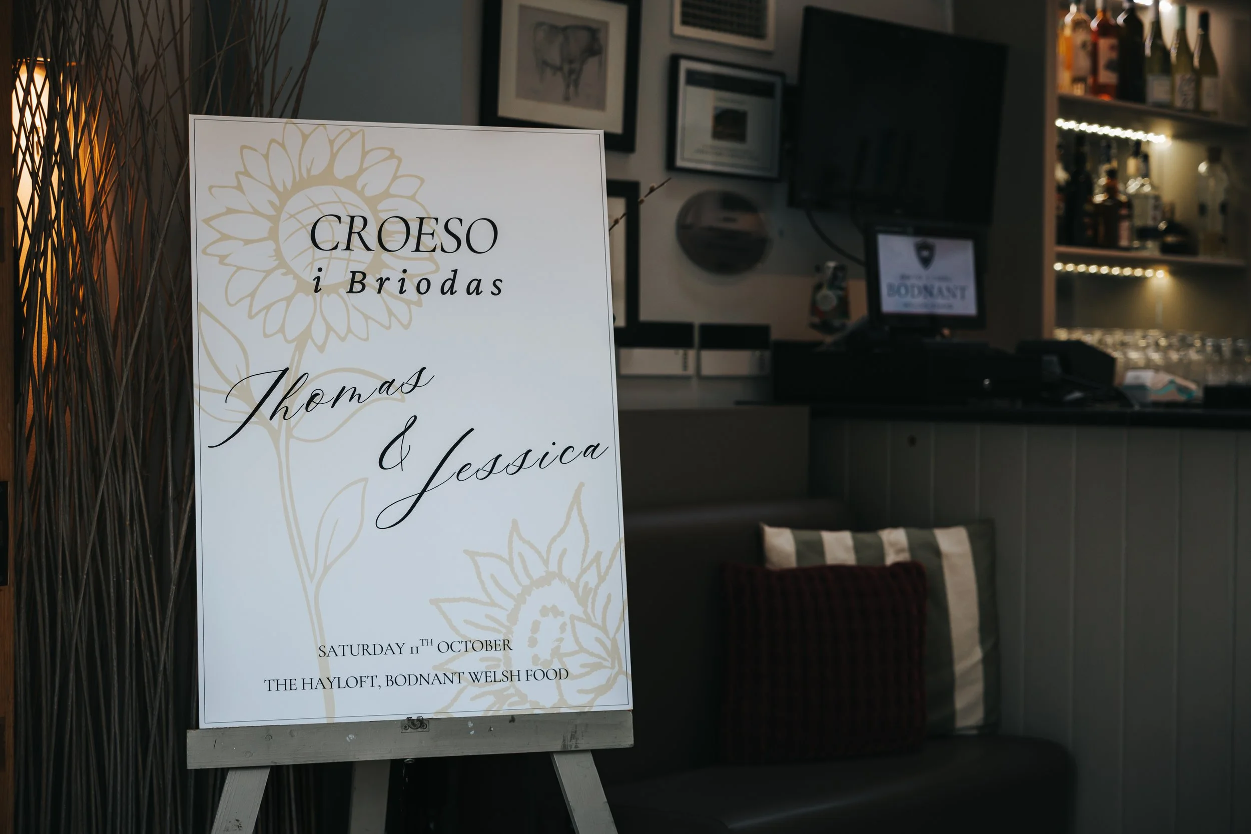 A white event sign with floral design announcing a wedding reception for Thomas and Jessica at The Hayloft, Bodnant Welsh Food on Saturday, October 11th.