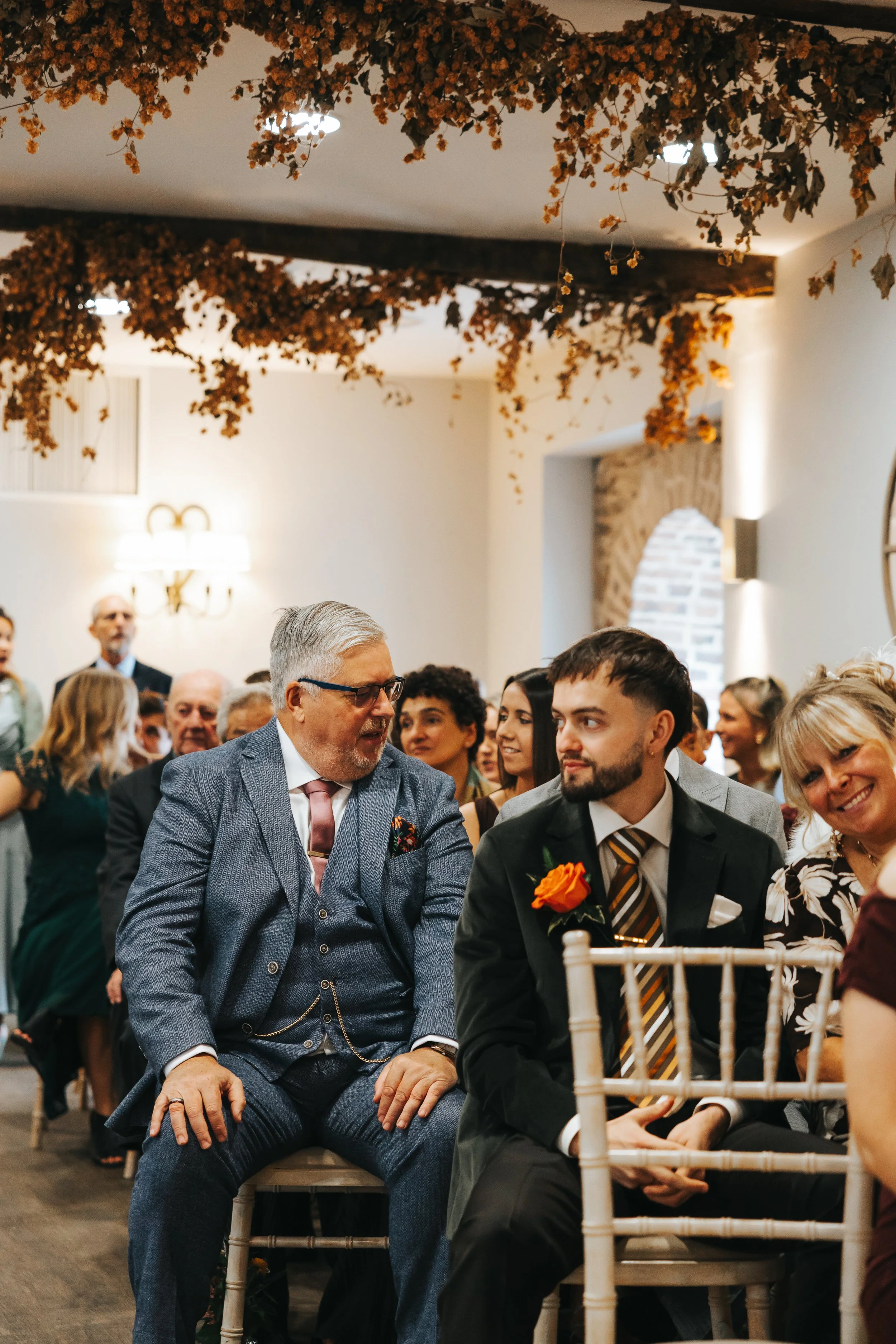 People attending a wedding ceremony, sitting on chairs, engaged in conversation, with some smiling. Decorations include hanging floral arrangements overhead.