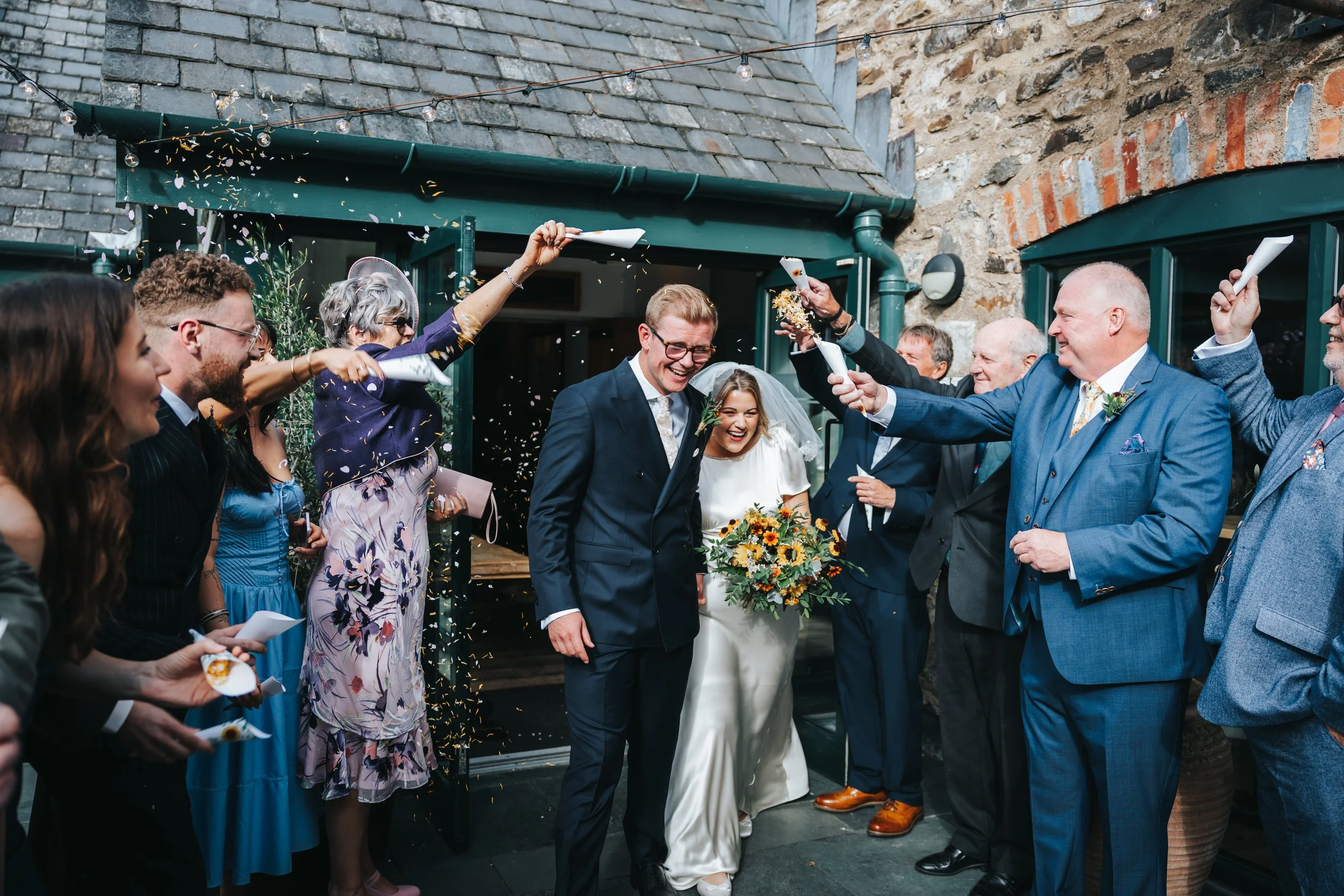 Bride and groom smiling as they walk through a shower of confetti thrown by friends and family outside a building.