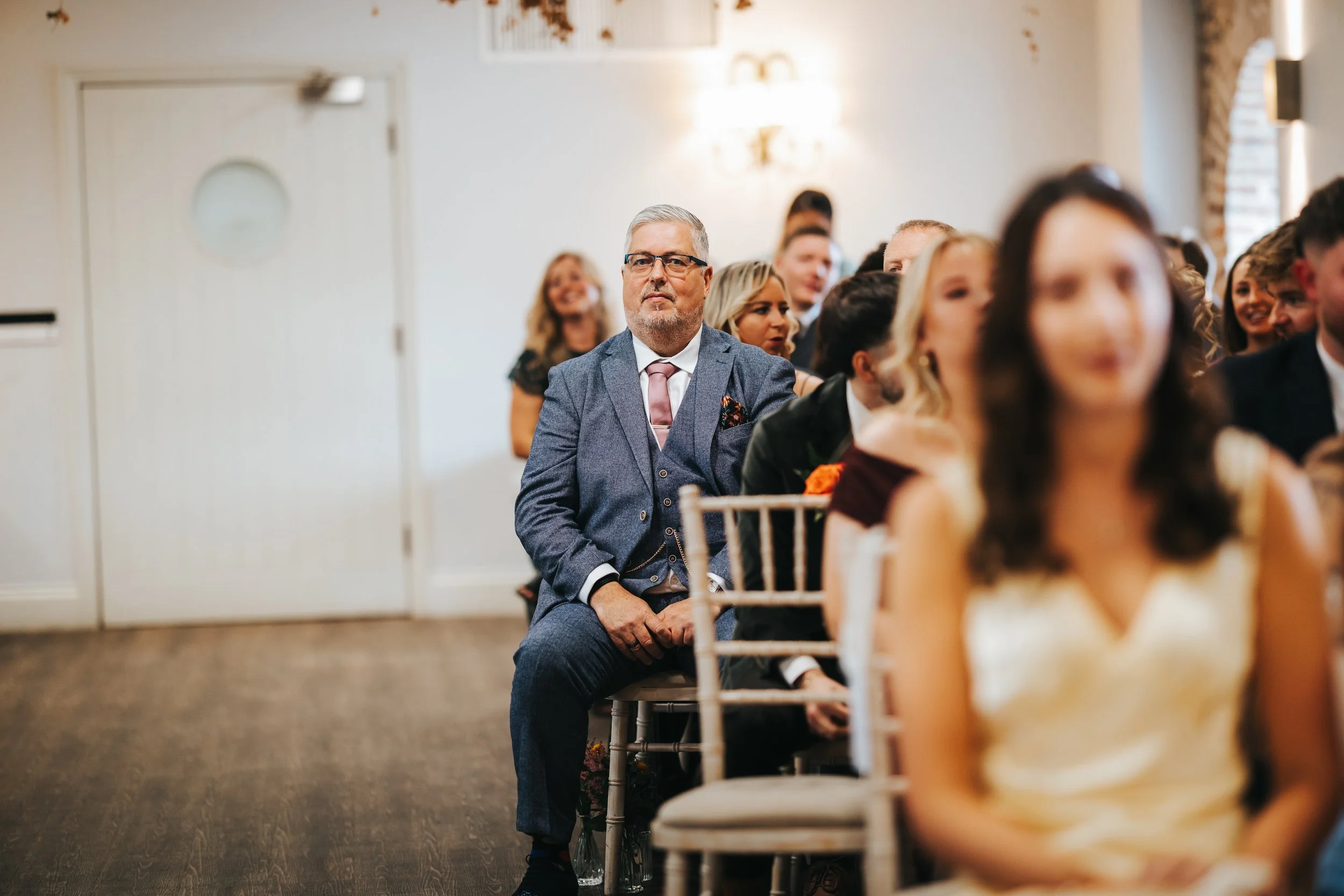 A group of formally dressed people attending a wedding ceremony, with a focus on a man in a gray suit and glasses sitting in the back, looking forward, while others around him sit and smile.