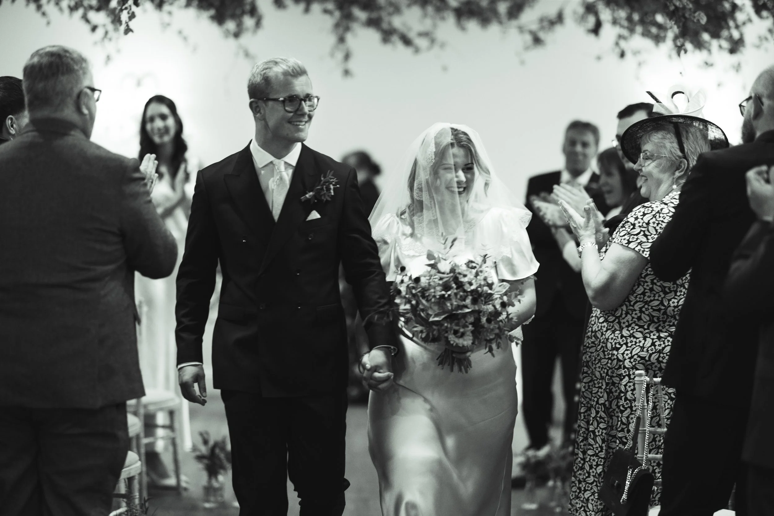 A bride and groom walking down the aisle holding hands, smiling, during a wedding ceremony, surrounded by clapping guests.