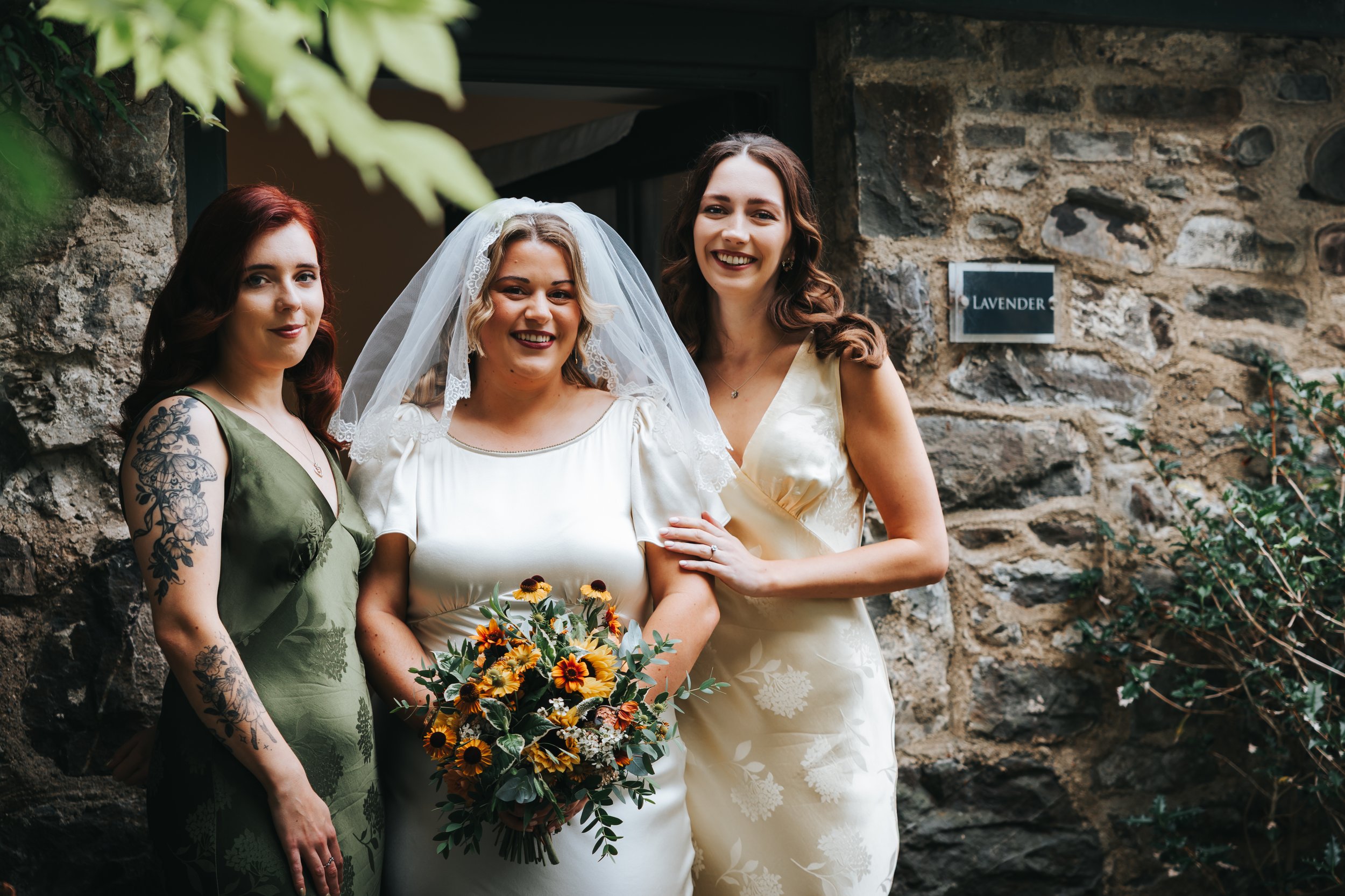 Three women standing outdoors in front of a stone wall, celebrating a wedding. One woman in the center wears a wedding dress, veil, and holds a bouquet of flowers. The woman on her left has red hair, a tattoo on her arm, and wears a green dress. The 