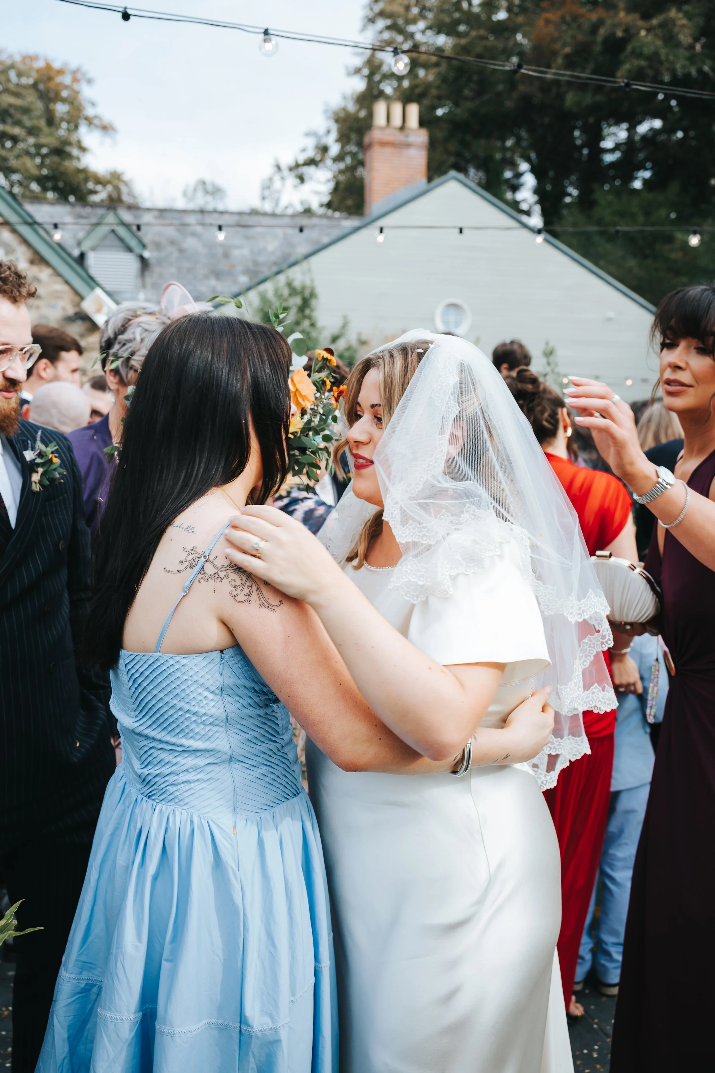 Two women, one in a wedding dress with a veil, and the other in a light blue dress, are embracing and looking at each other at an outdoor wedding reception with guests and string lights overhead.