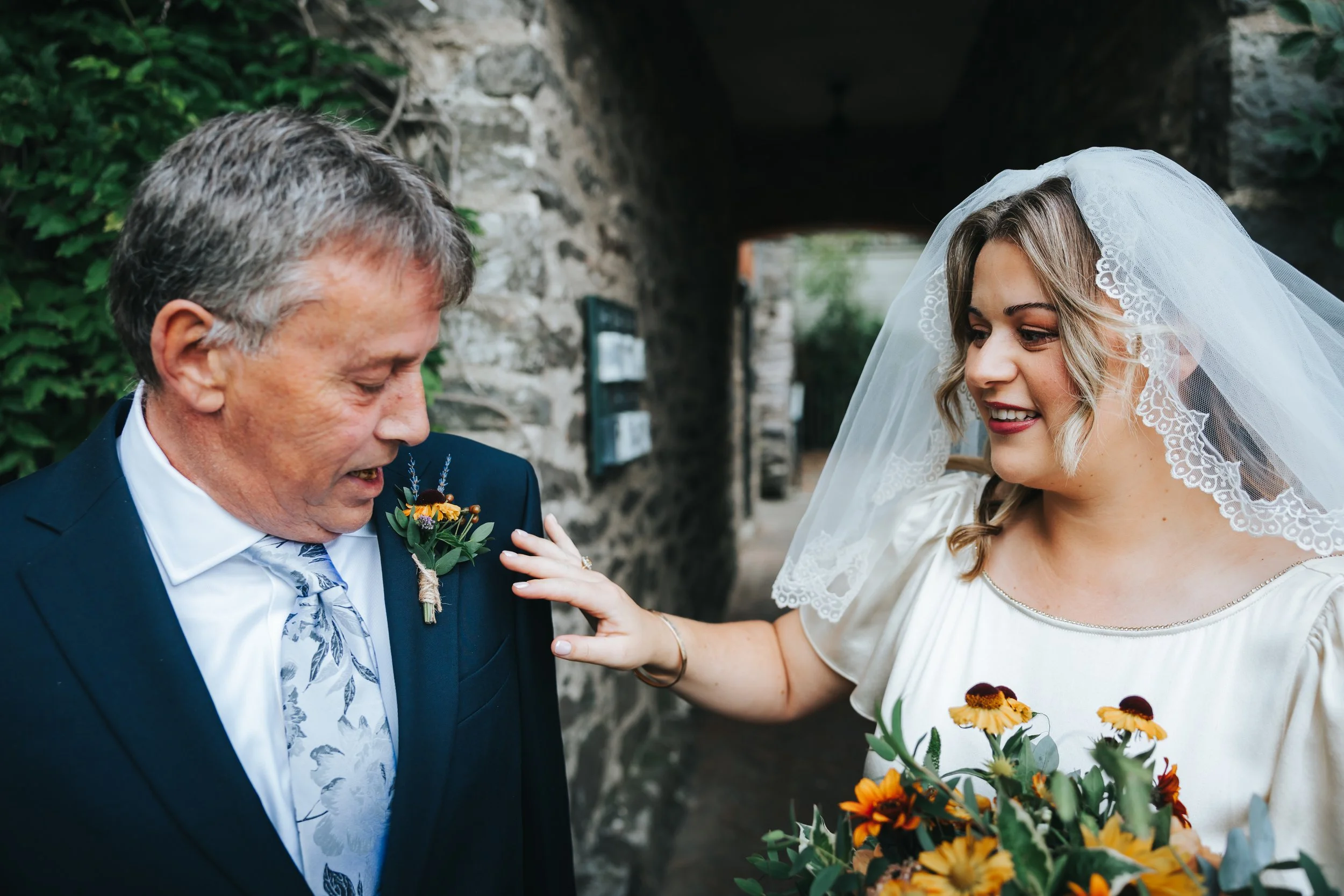 A bride in a white dress and veil touching an older man's shoulder, who is wearing a dark suit and tie, both smiling outdoors next to a stone wall holding a bouquet of yellow flowers.