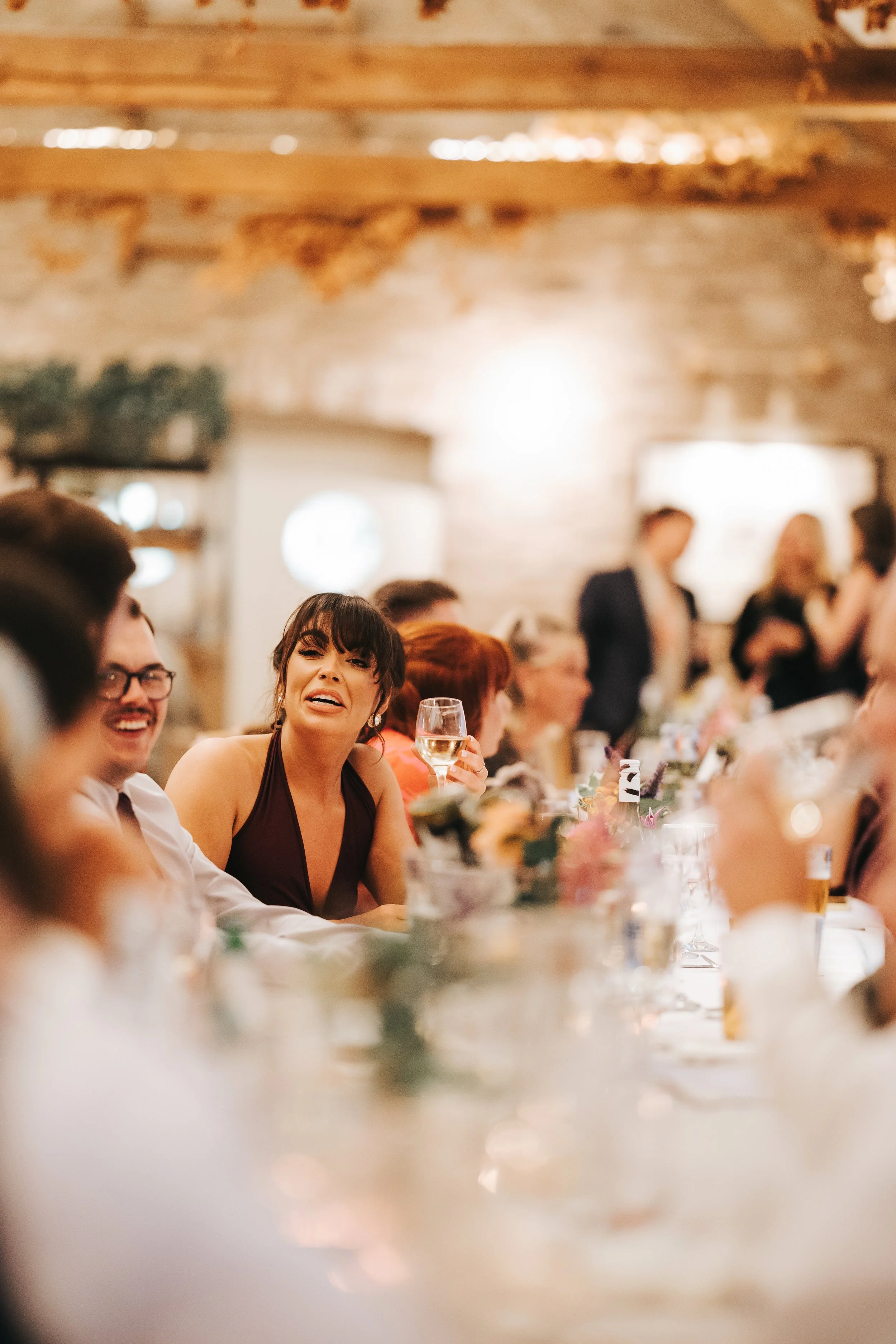 People sitting at a decorated banquet table during a celebration, with a woman in a dark dress holding a glass of wine, smiling and engaging with others in a warmly lit rustic setting.